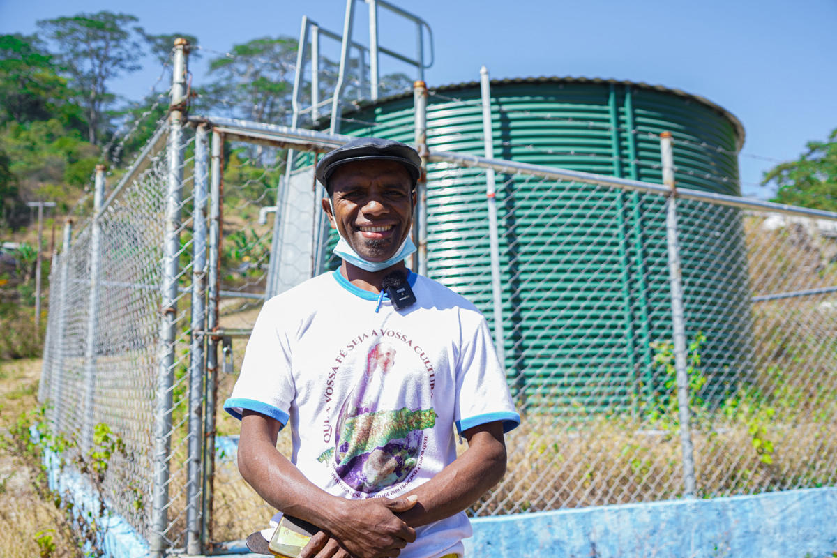 Man in a light shirt stands by a chain-link fence with a large green water tank behind.