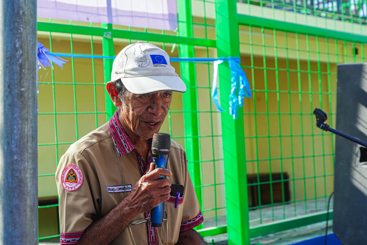 Scout in khaki uniform speaks into a microphone outdoors beside a bright green fence.