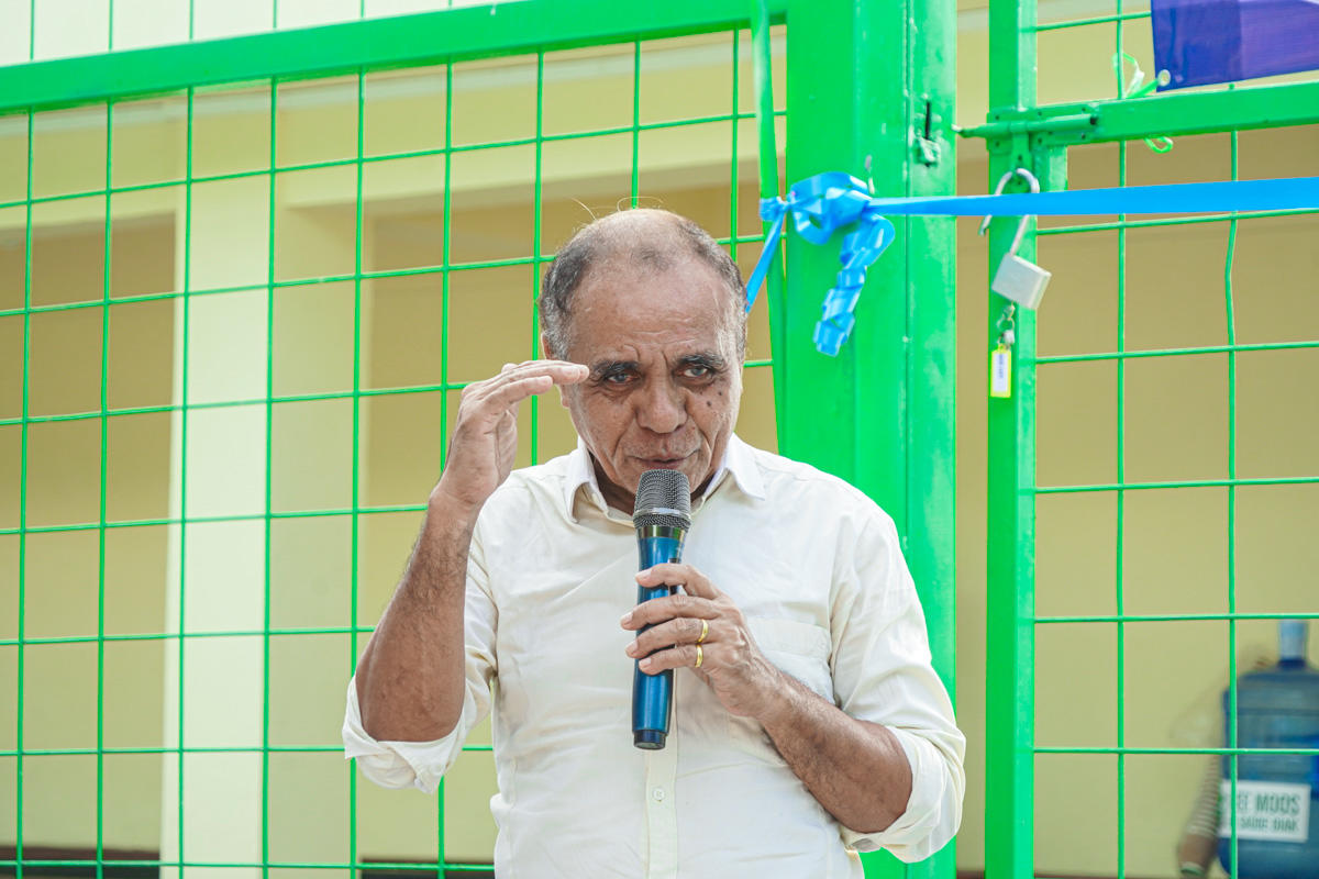 Man in a white shirt speaks into a microphone beside a bright green fence.