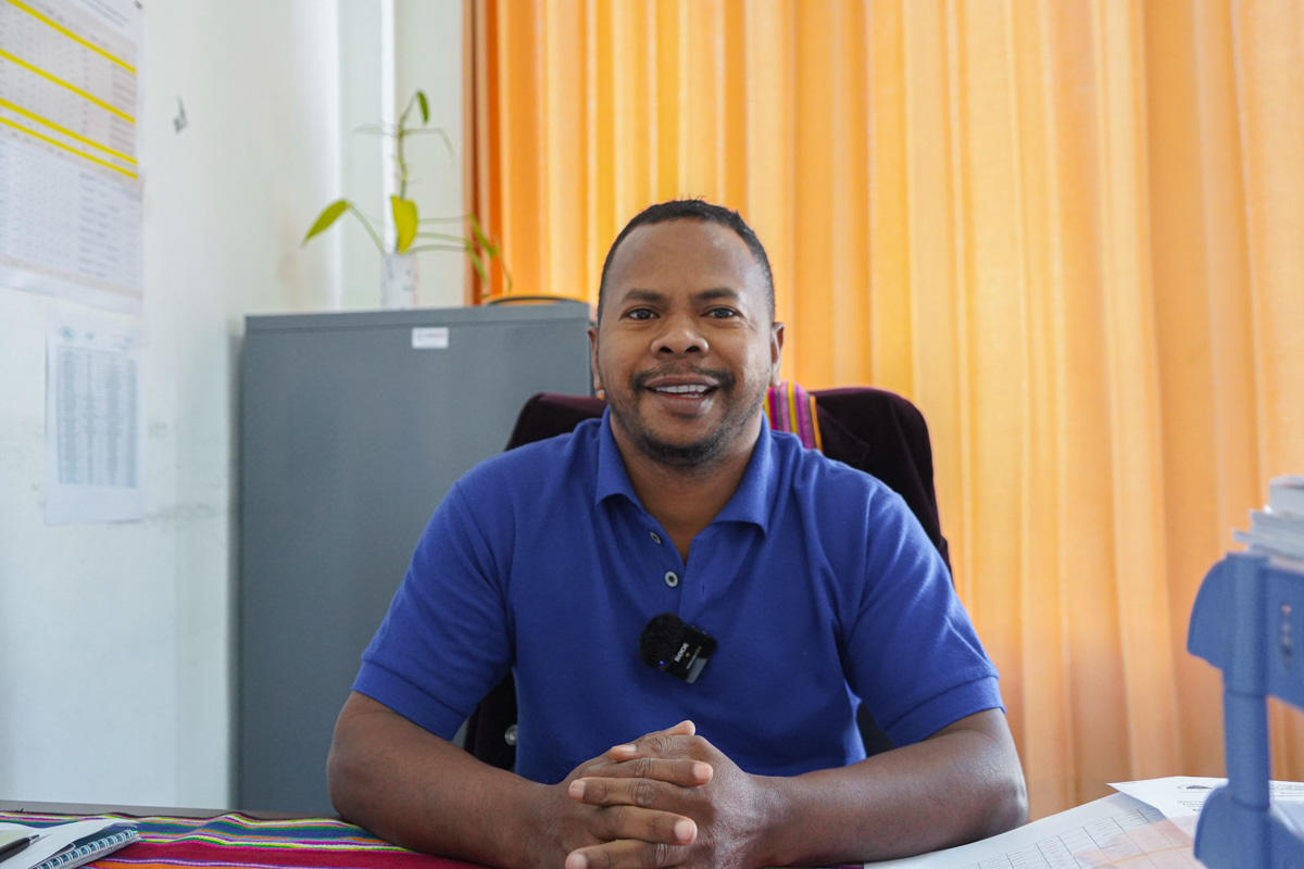 Person in blue polo sits at a desk in an office with orange curtains behind.