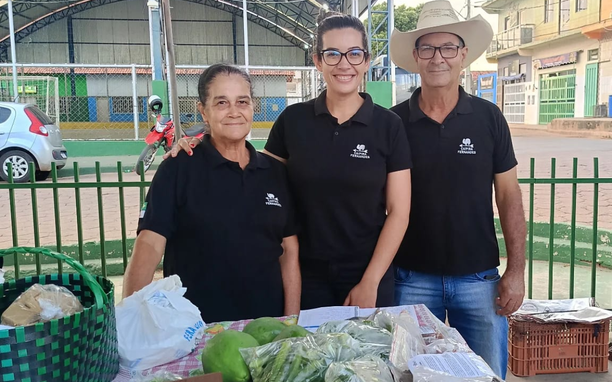 Photo: Four smiling volunteers in black polo shirts stand behind a table of vegetables outdoors.