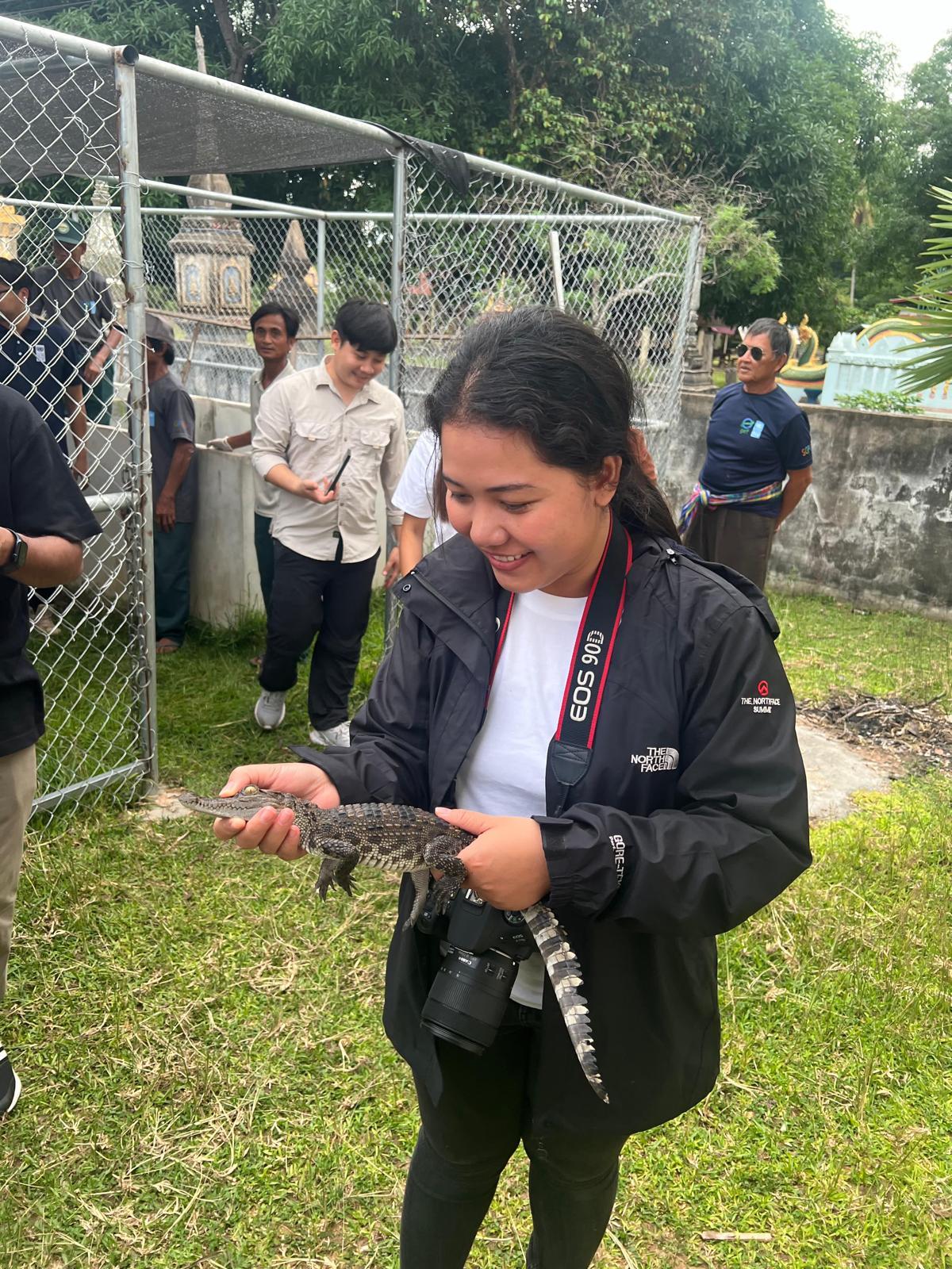 Photograph of a person in a black jacket with a camera, standing on grass near a chain-link fence.