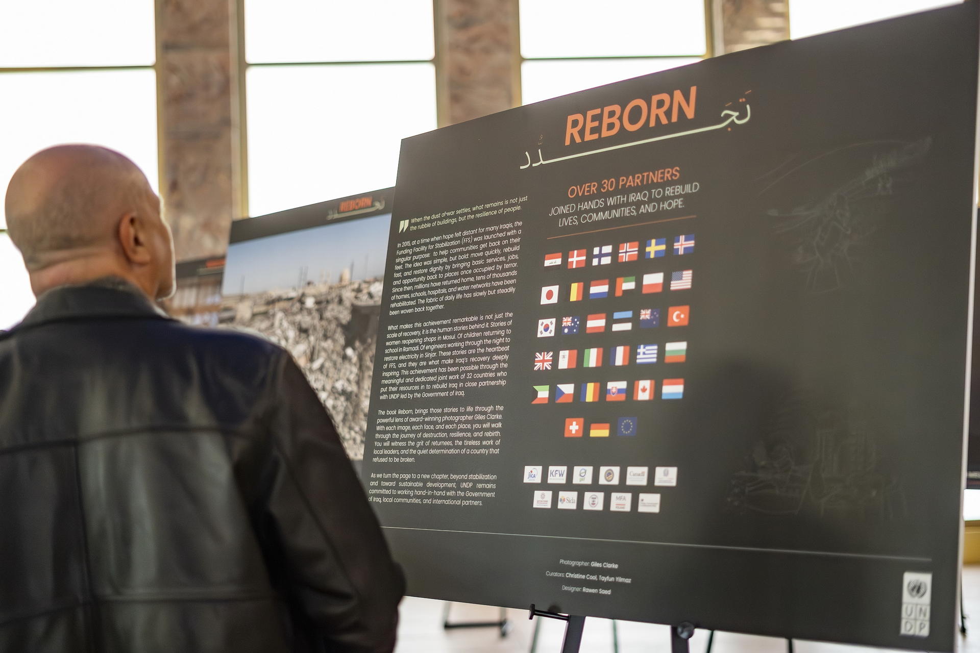 Man in a leather jacket examines a large display board titled REBORN with many thumbnails on screen.