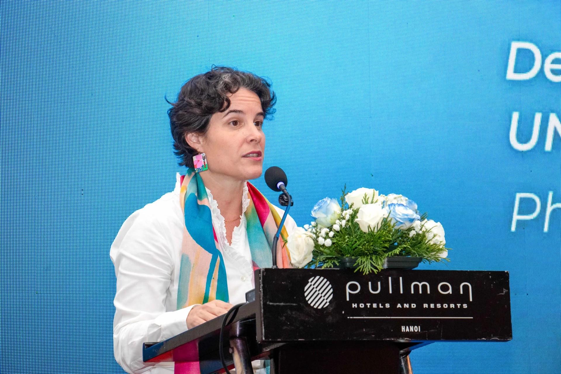 Speaker at a Pullman podium, blue backdrop, white coat with teal scarf, flowers on lectern.