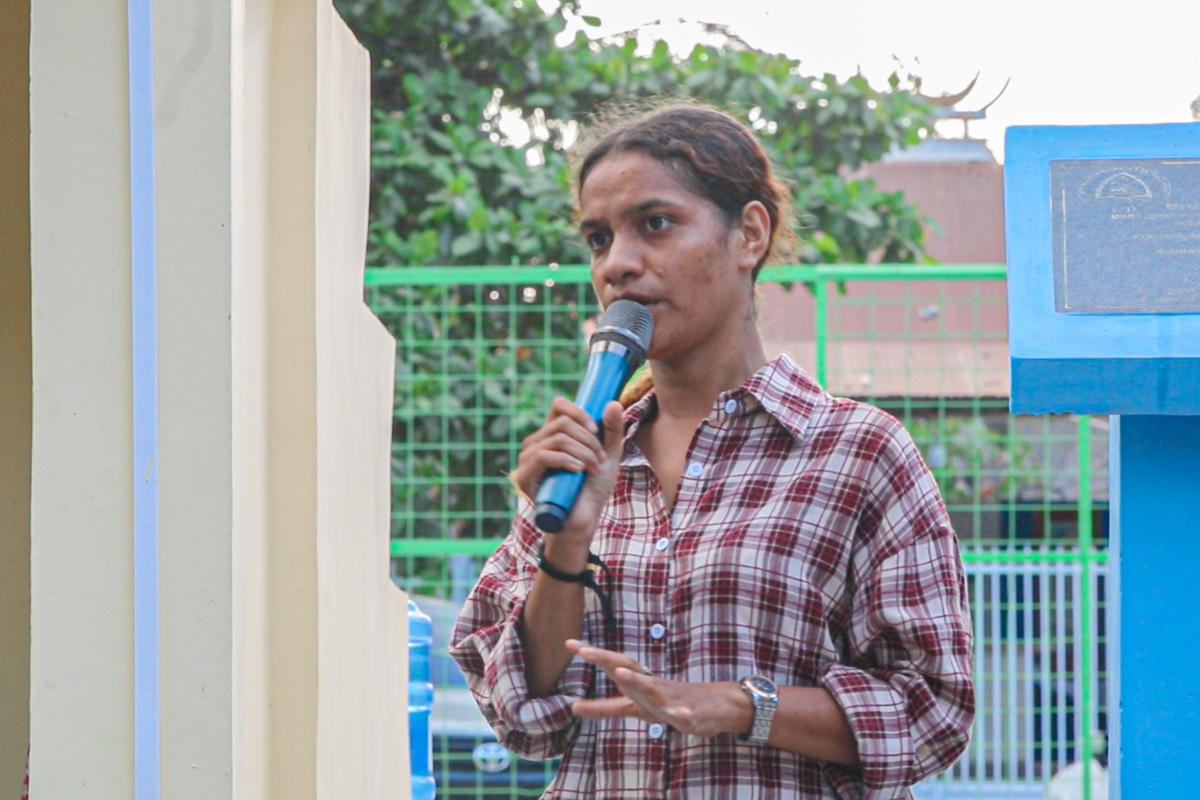 Man in red plaid shirt speaks into a microphone outdoors beside a blue sign and green fence.