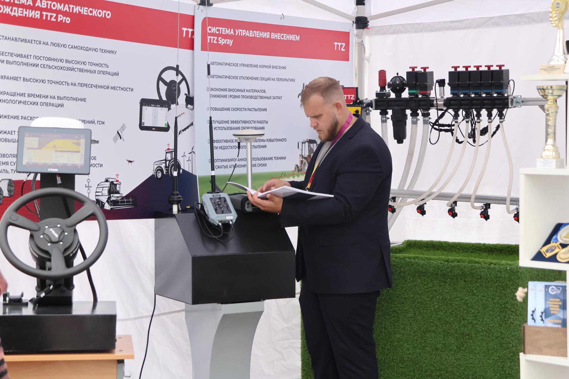 Man in a suit examines a device at a trade show booth with machinery and banners.