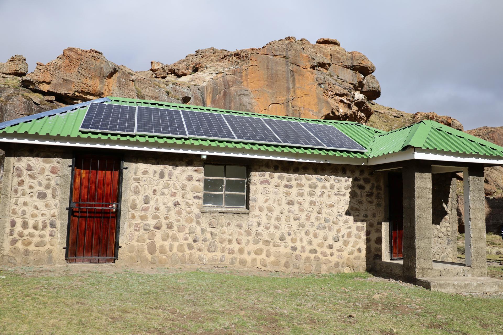 Stone cottage with green roof and solar panels, red door, beneath rocky cliffs.
