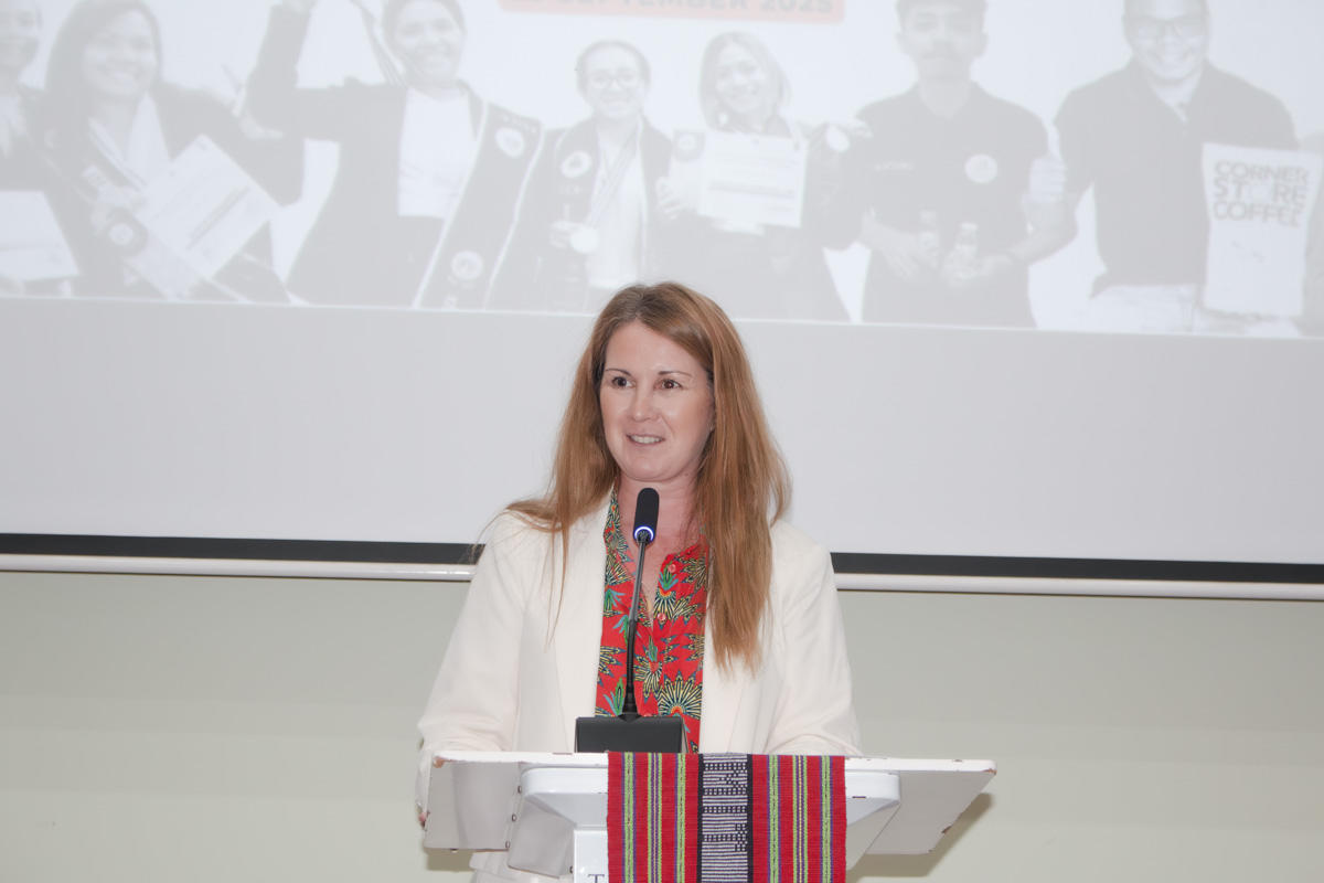 Presenter at podium in white jacket and colorful scarf, projected crowd on screen behind.