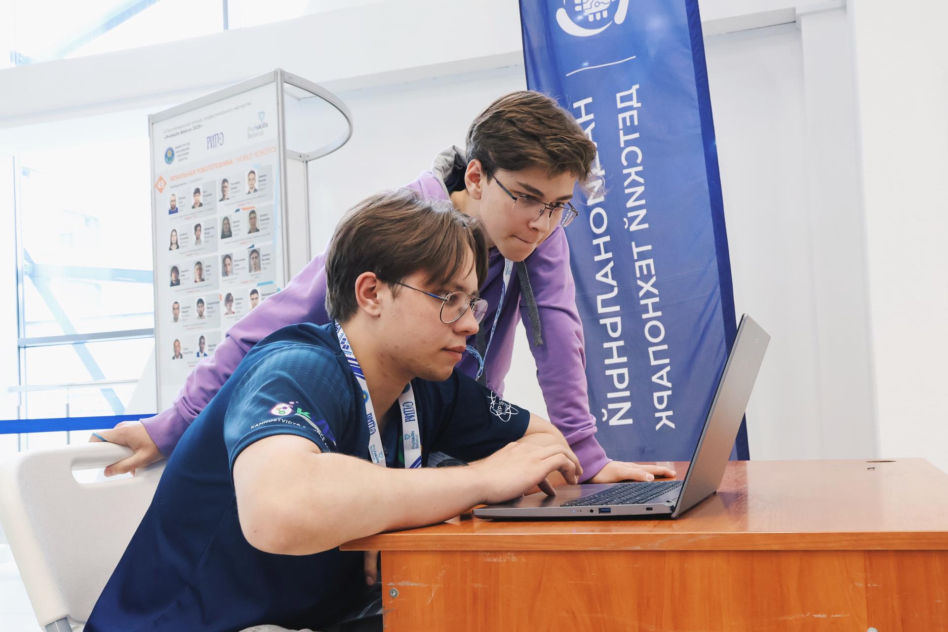 Two people lean over a wooden desk with a laptop, blue banner in a bright room.