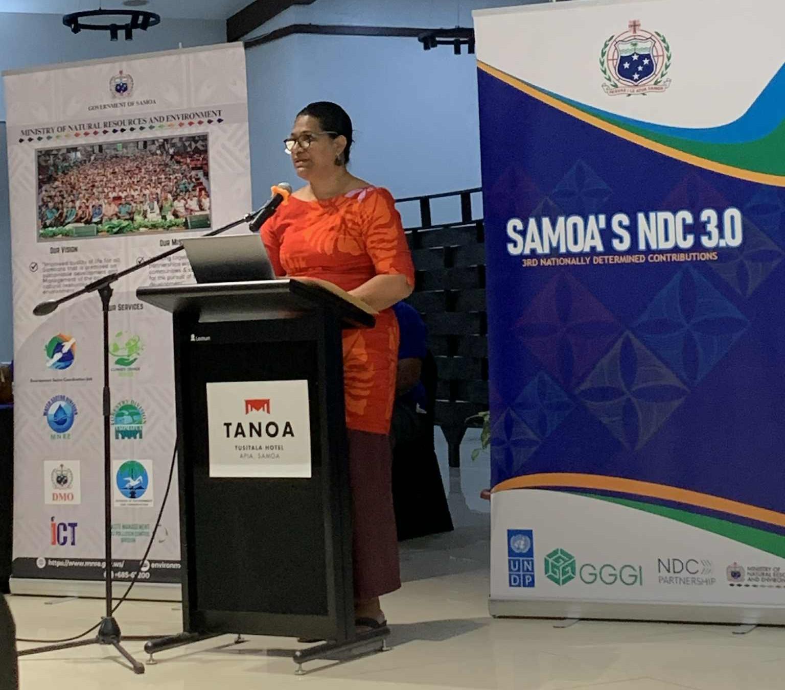 Woman in an orange dress speaks at a podium on a stage, flanked by banners including Samoa's NDC 3.0.