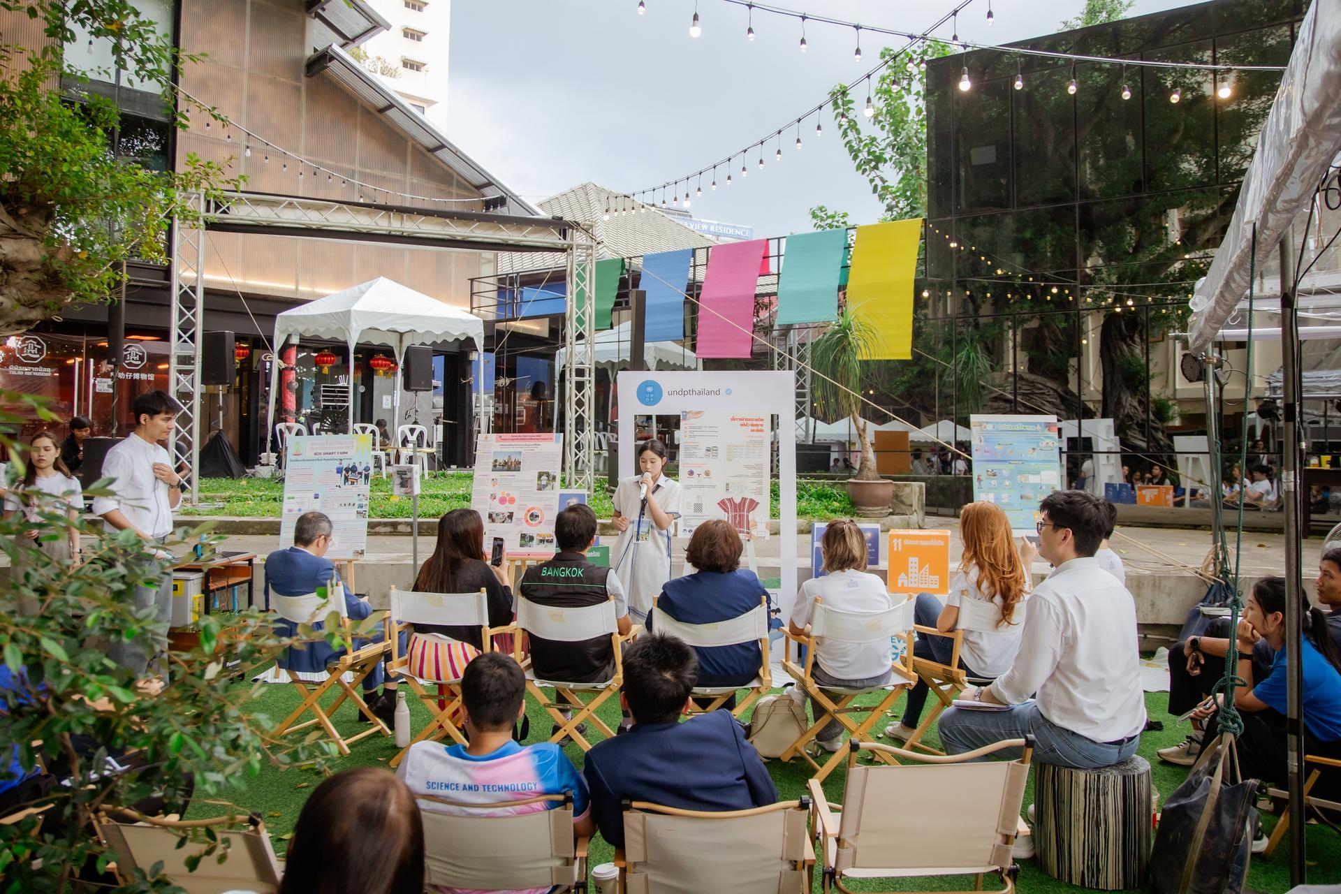 Outdoor community event with audience watching a performer on a small stage and colorful banners.