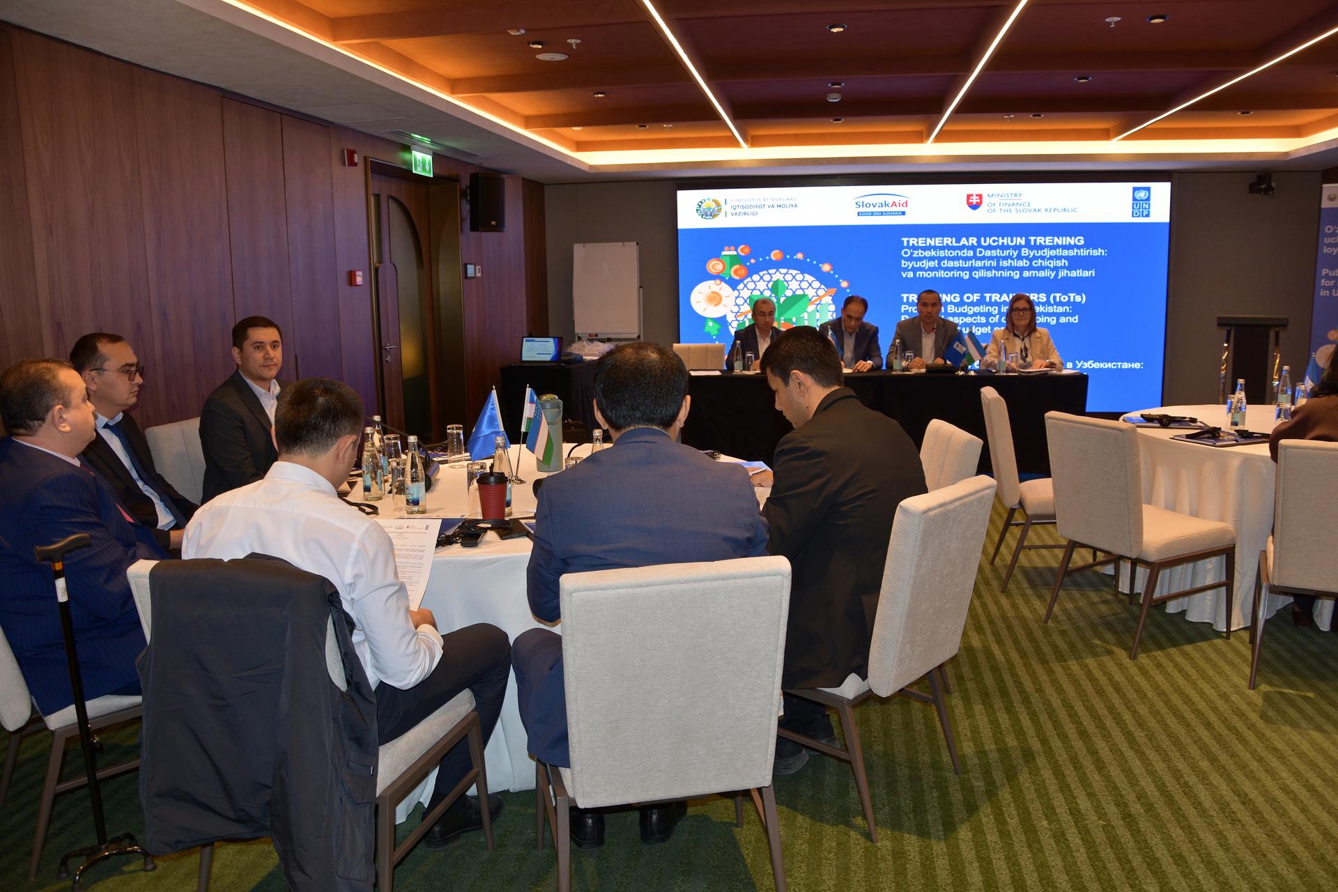 Attendees at a business conference sit around tables; presentation screen on front wall.