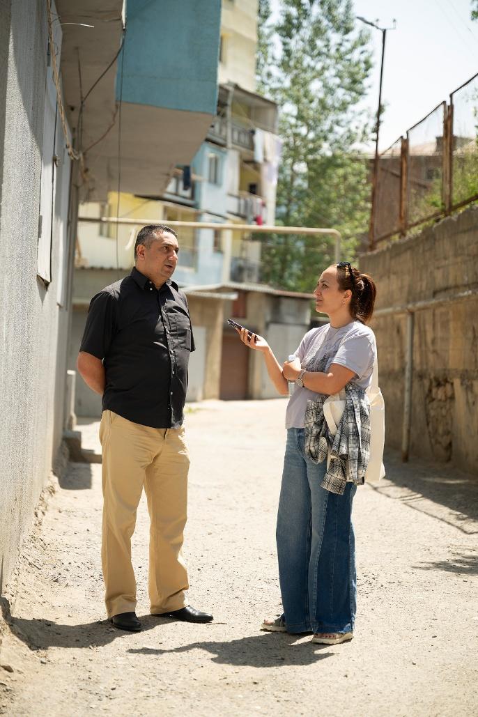 Man and woman talk in a sunlit alley between beige buildings.