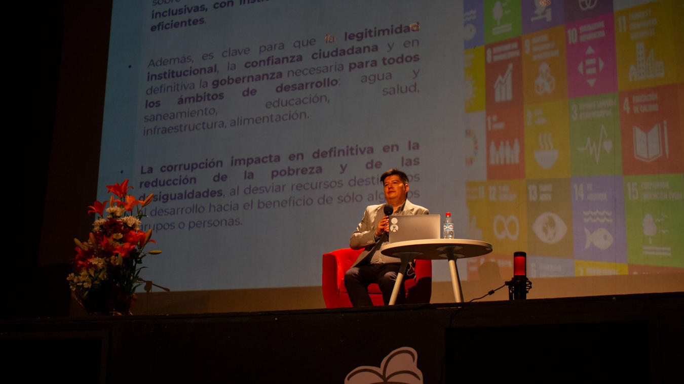 Photograph of a speaker at a podium on stage, with a floral arrangement on the left and a colorful screen behind.