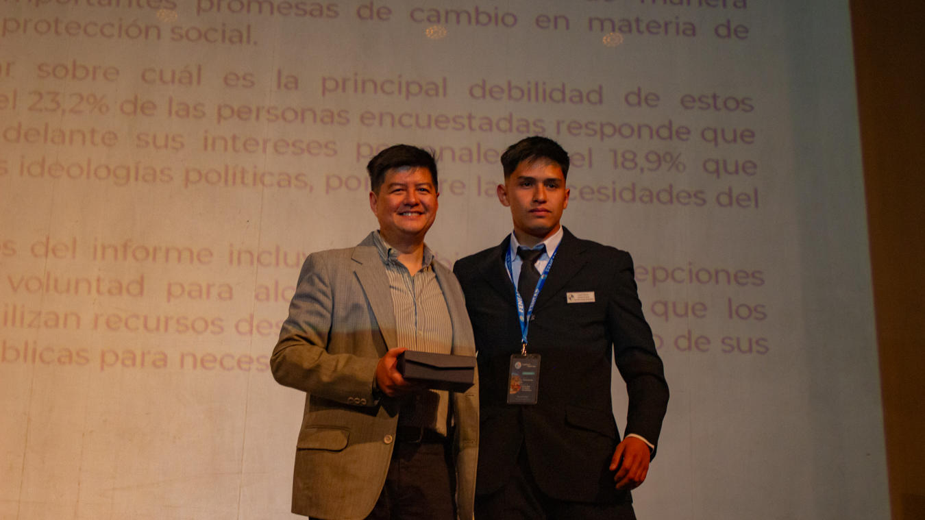 Photograph: two men in suits on stage, one receiving an award, projection screen with text behind.