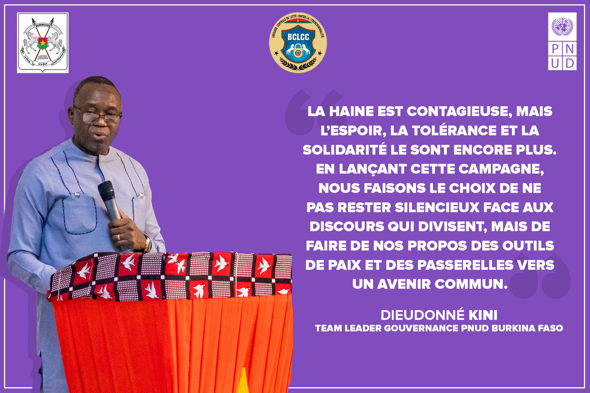 Photograph of a man in a blue shirt speaking at a podium on a purple stage with banners and logos.