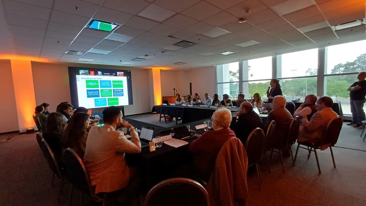 Grupo de personas alrededor de una mesa de conferencias en forma de U; pantalla a la izquierda, grandes ventanales al fondo