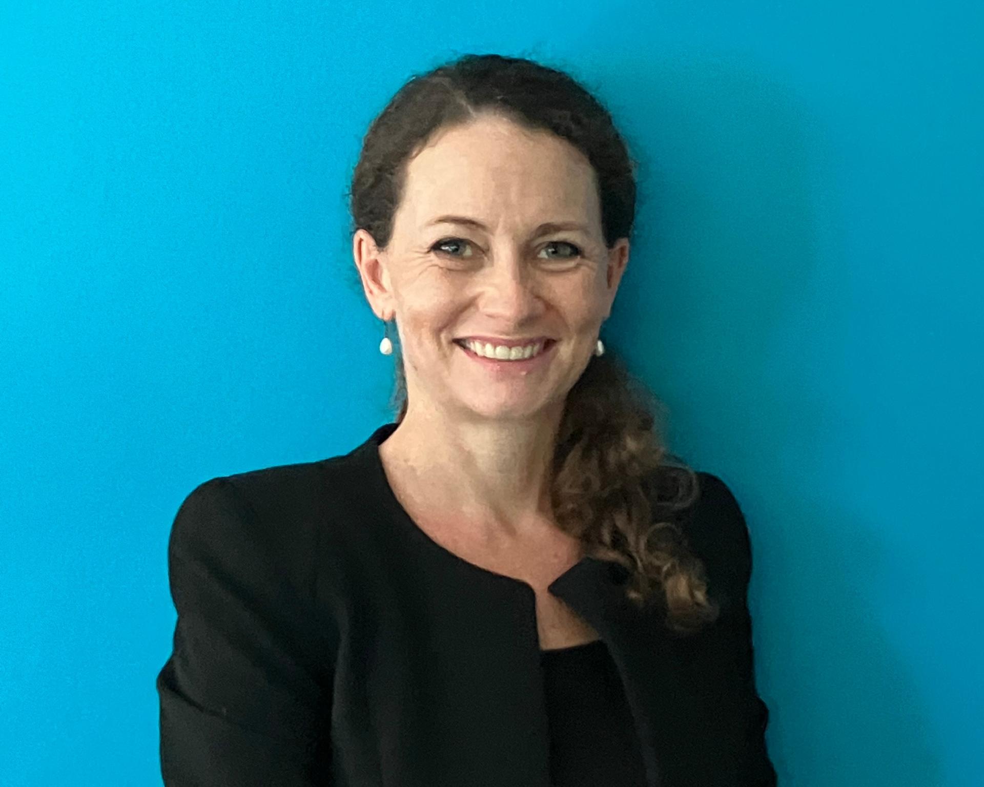 Photograph of a woman with a ponytail in a black blazer against a bright blue wall.