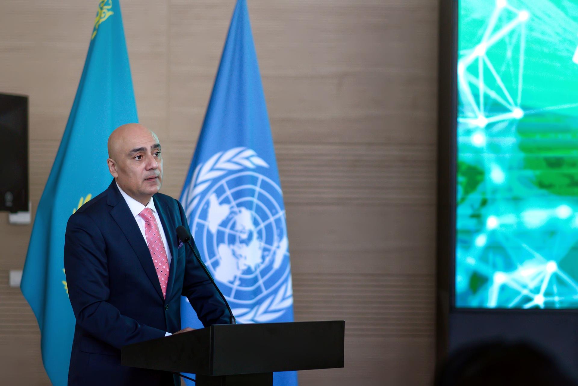 Speaker at podium with UN flag and blue flag behind him; green digital screen to the right.