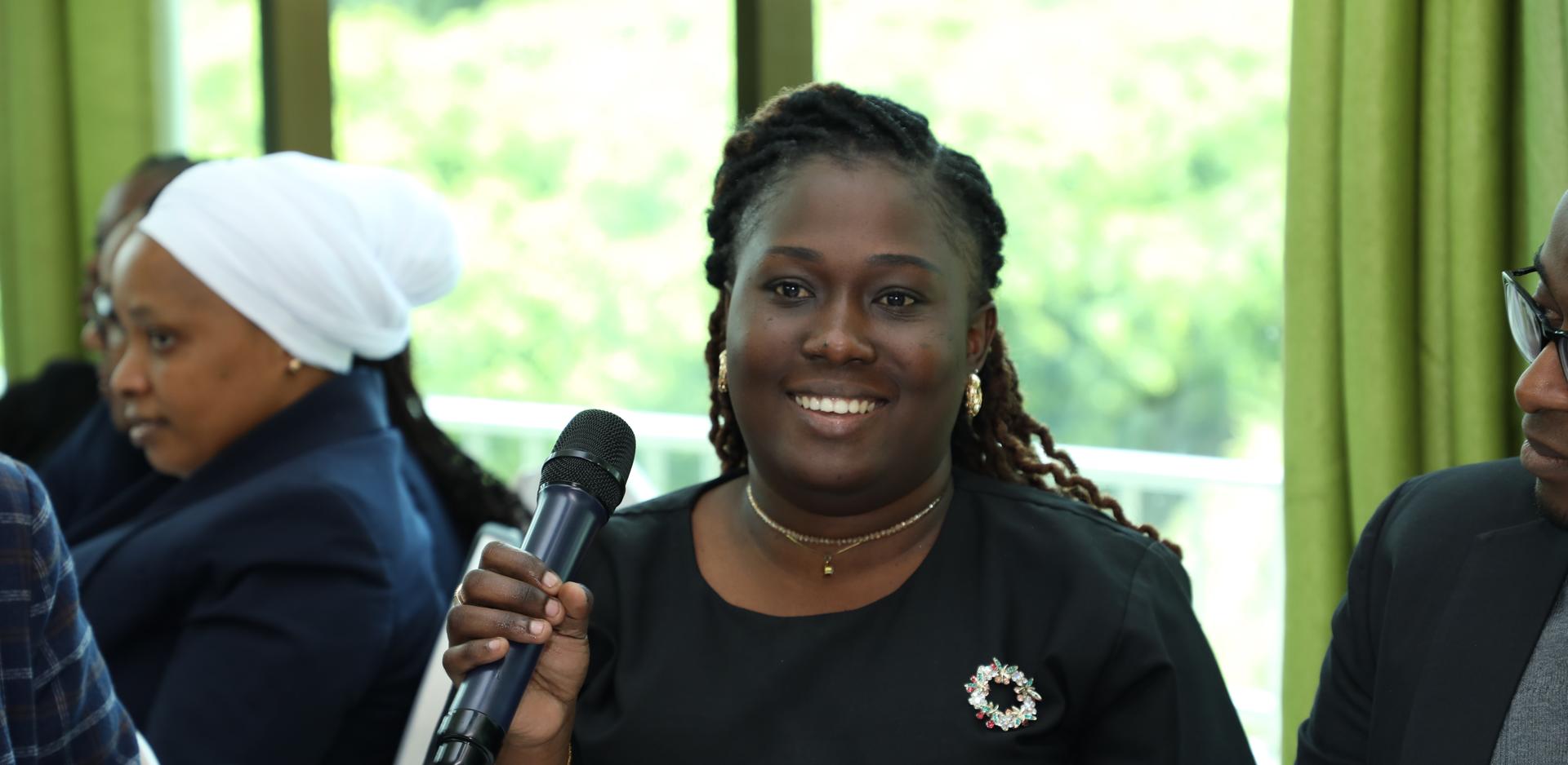 Speaker in black top holds a microphone, with people surrounding her and green landscape visible through the window behind her.