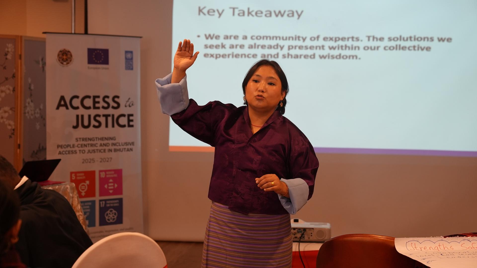 Presenter in maroon top and striped skirt gesturing at a slide; left banner reads ACCESS & JUSTICE.
