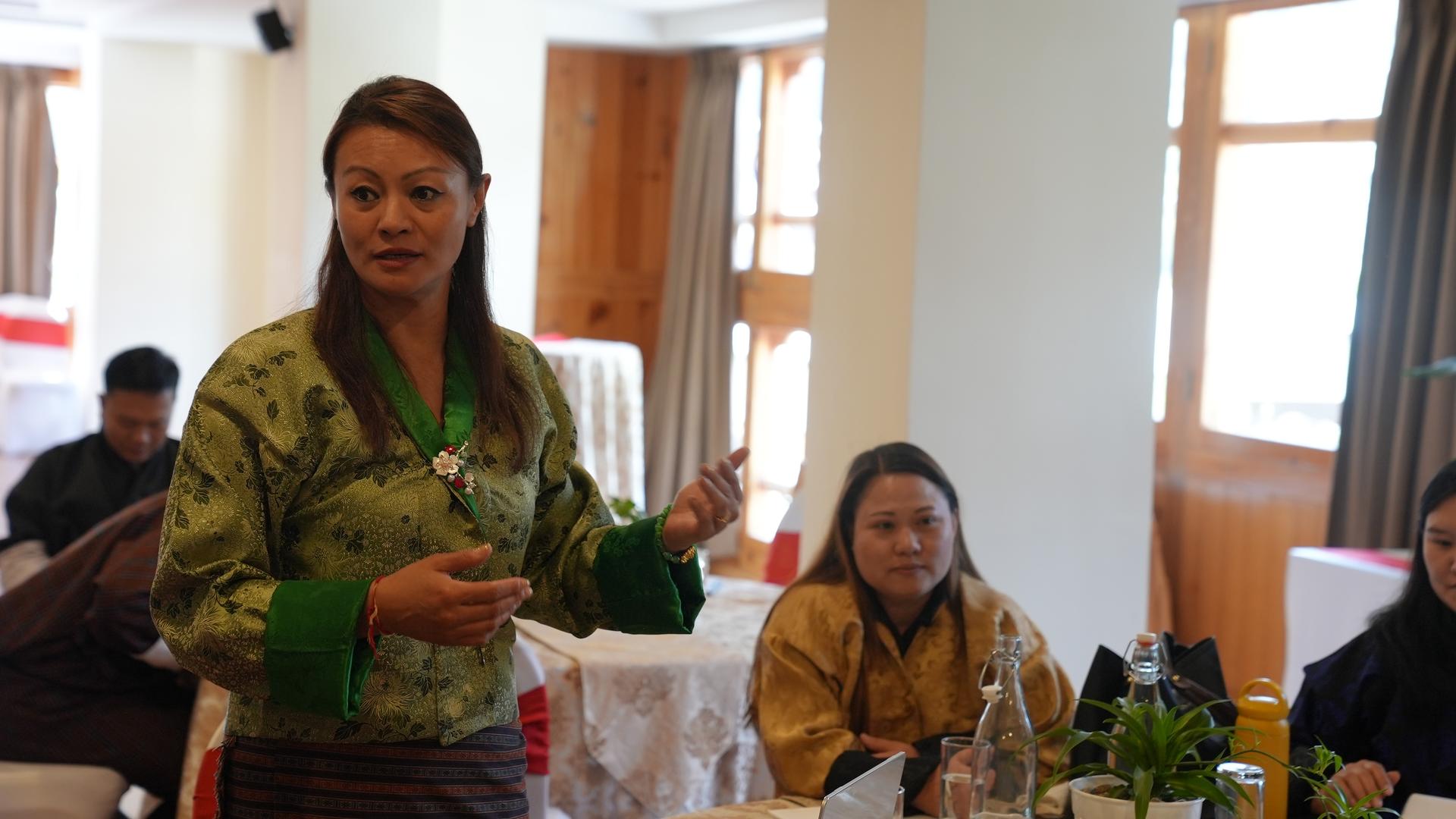 Group of people in a bright meeting room; person in green patterned jacket speaks at the table.