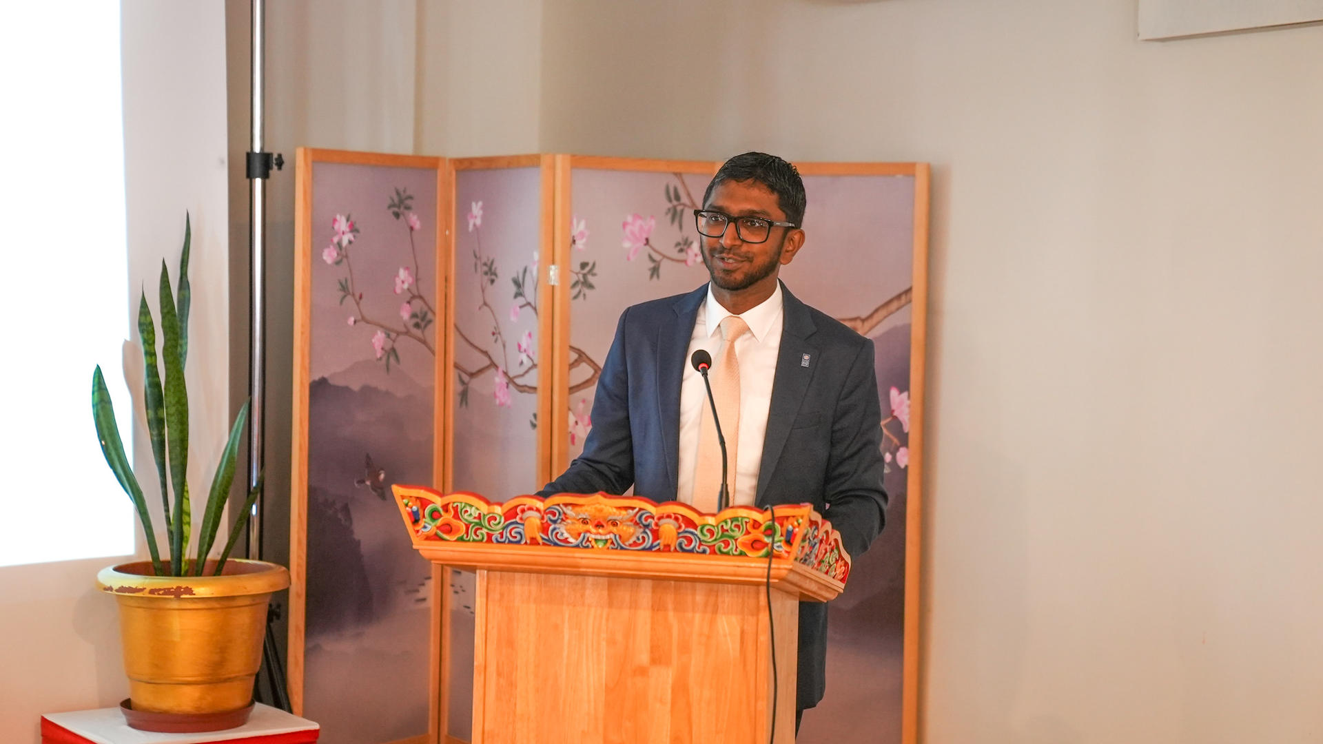 Man in a suit delivers a speech at a wooden podium, with a potted plant and folding screen behind.