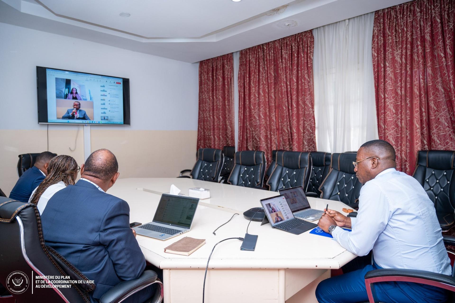 Business meeting in a conference room; team with laptops; video call on wall monitor.