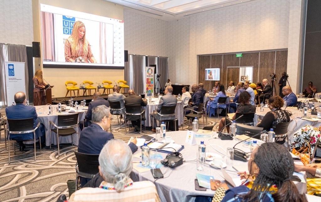 Panel discussion at a conference in a ballroom; speaker on screen, audience at round tables.
