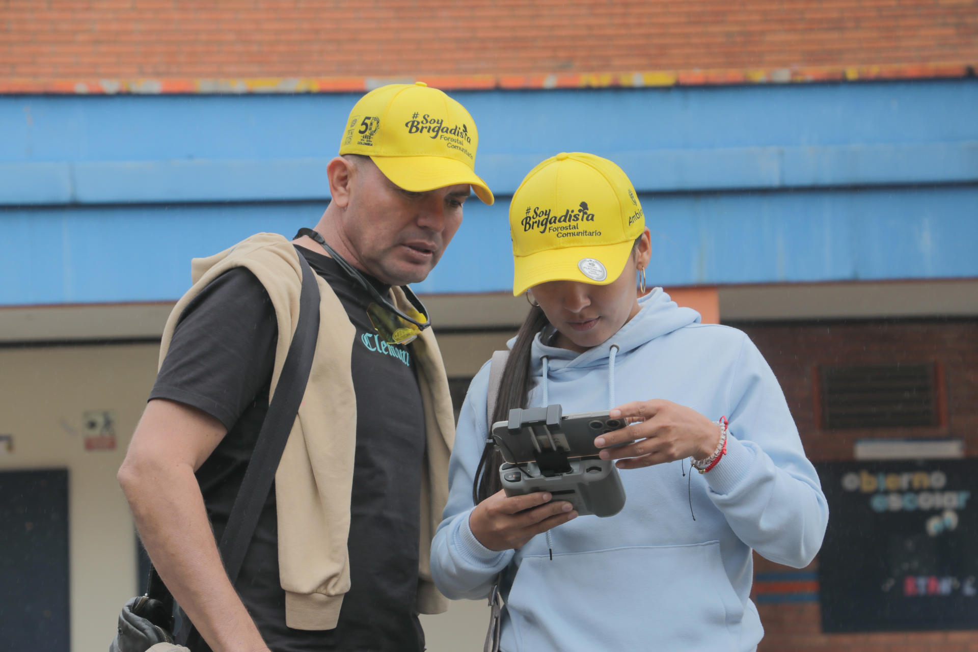 Two men outdoors; one in a yellow helmet and tan vest, the other in a blue hoodie with a remote.