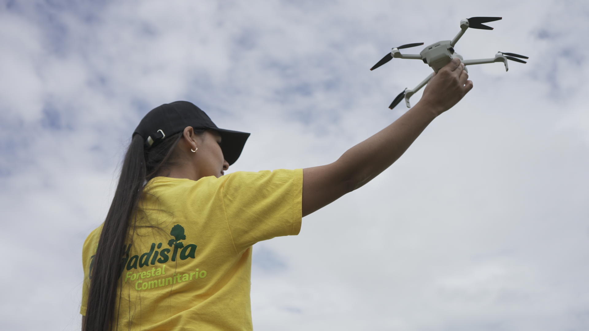Photograph of a person in a yellow shirt holding a quadcopter drone against a cloudy sky.