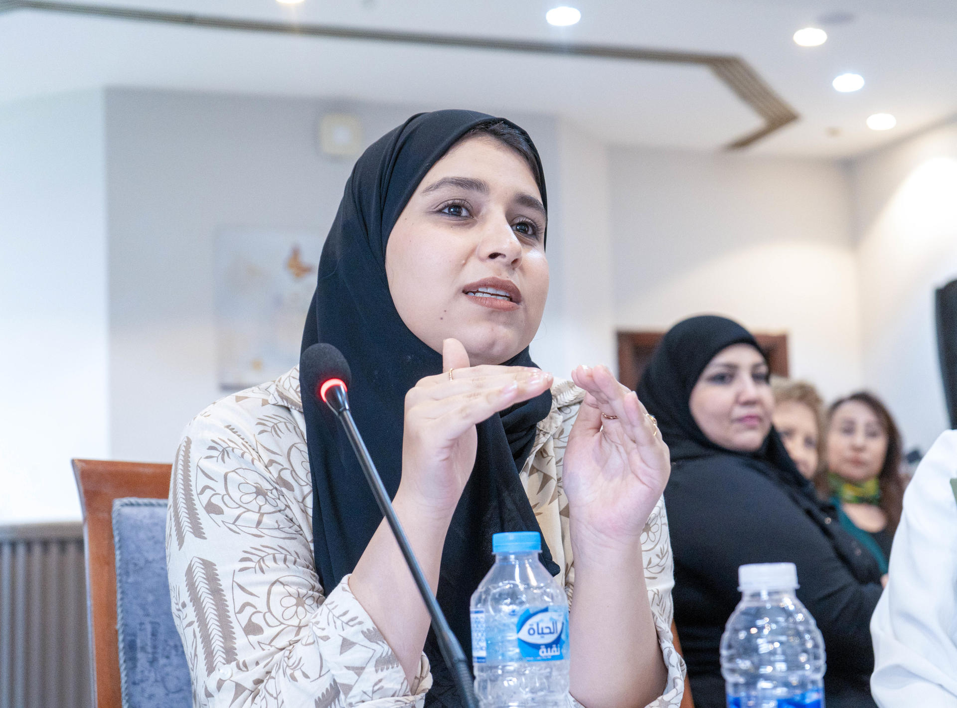 Panelist with hands near mouth at a conference table, microphone and water bottles.