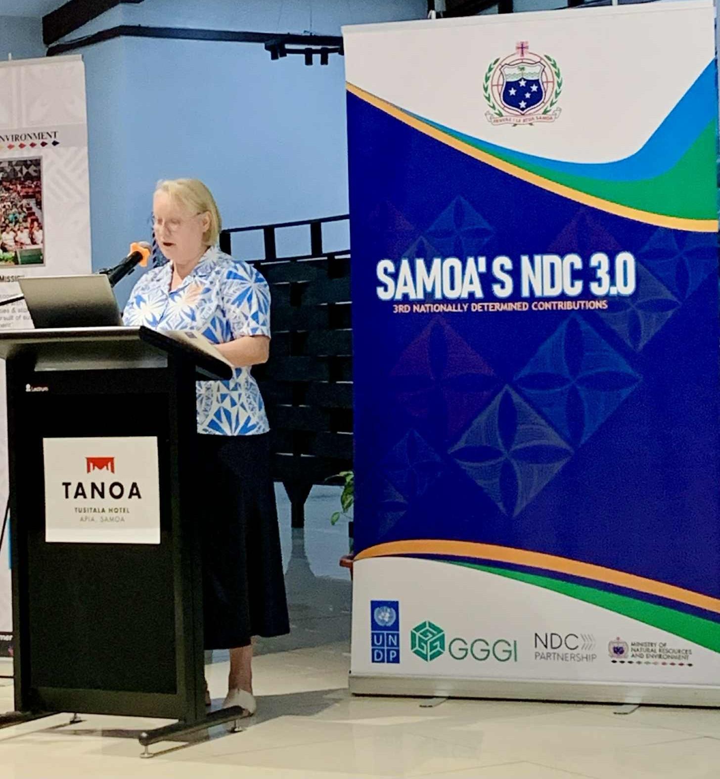 Woman speaker at podium with a Tanoa sign beside a blue Samoa's NDC 3.0 banner.