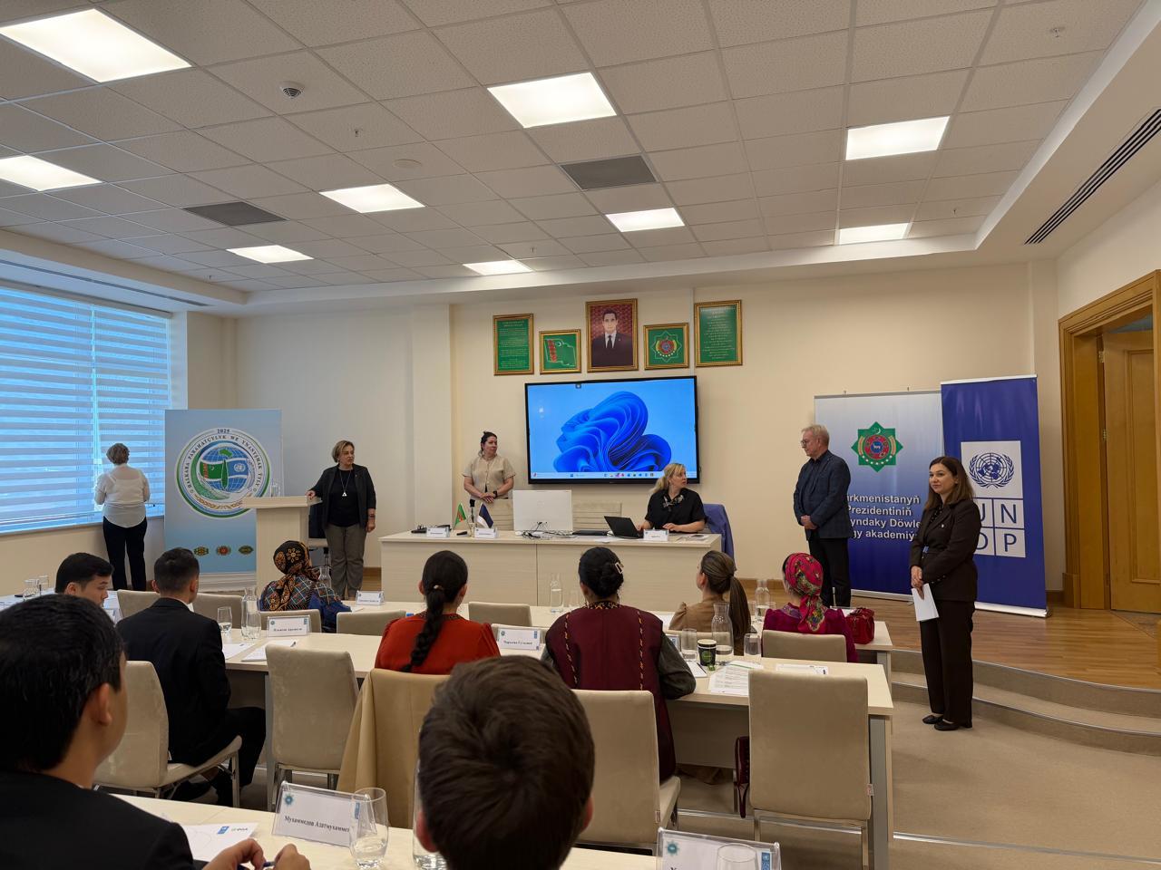 Photograph: conference room with panelists at a long table, seated audience, banners, and screen.