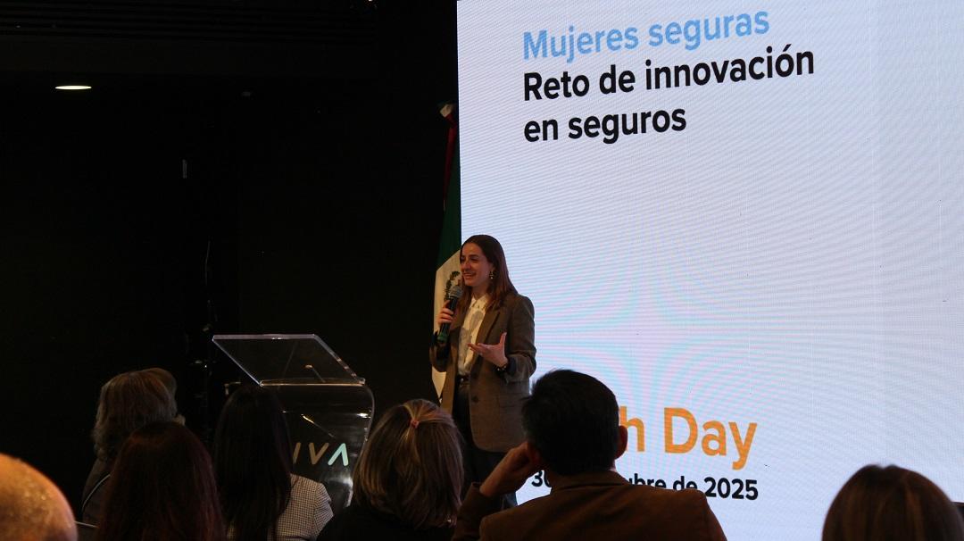 Female presenter on stage at a conference with a Spanish slide behind her, audience in foreground.