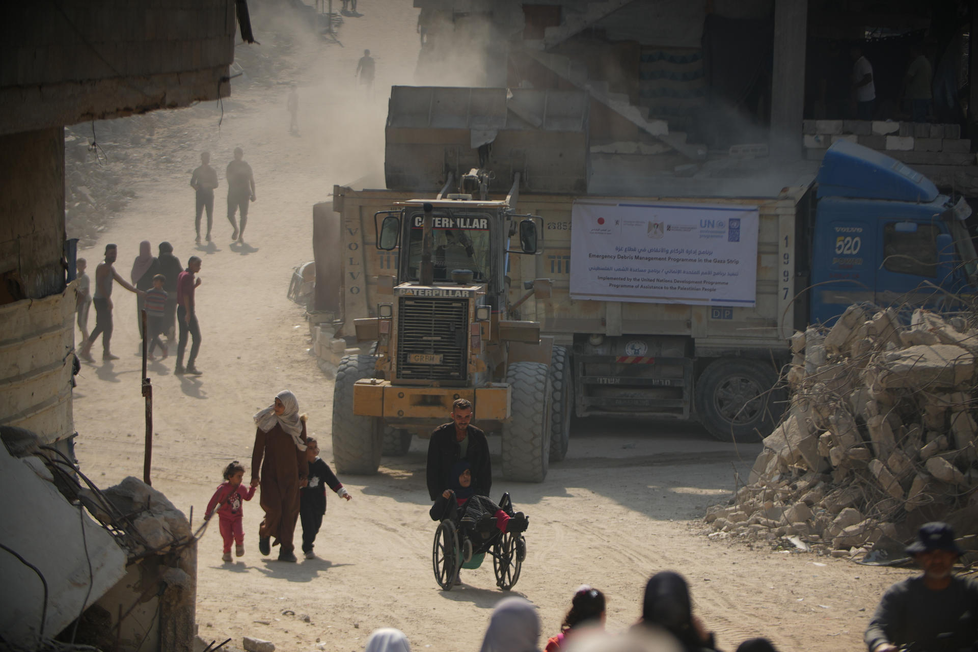 Dusty construction street with a yellow bulldozer, pedestrians, a motorbike, and a blue container.