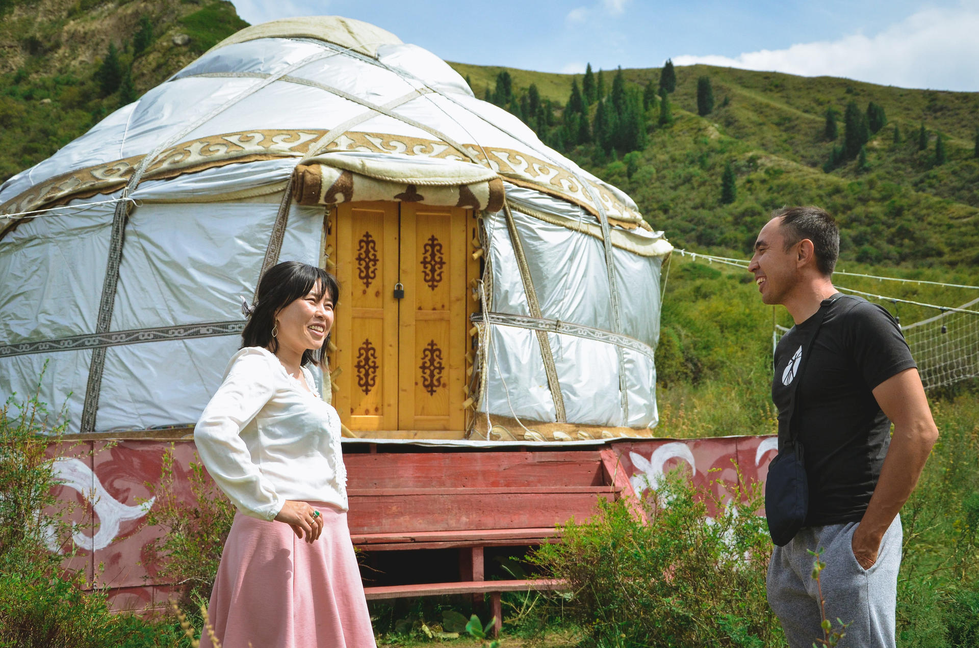 Photograph of a woman in a pink skirt and white top talking to a man beside a white yurt.