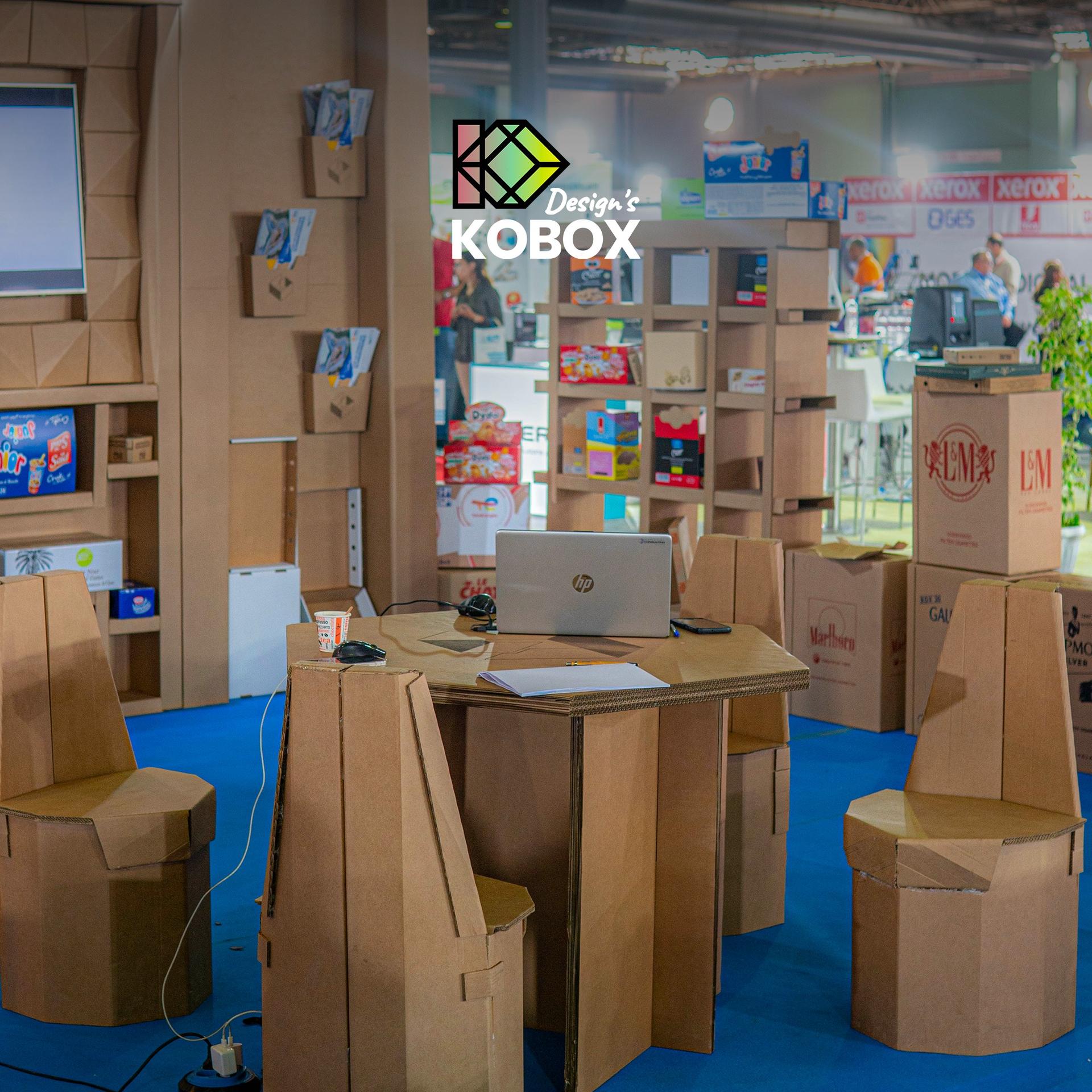 KOBOX logo visible at a booth with blue carpet, cardboard furniture, and laptop.