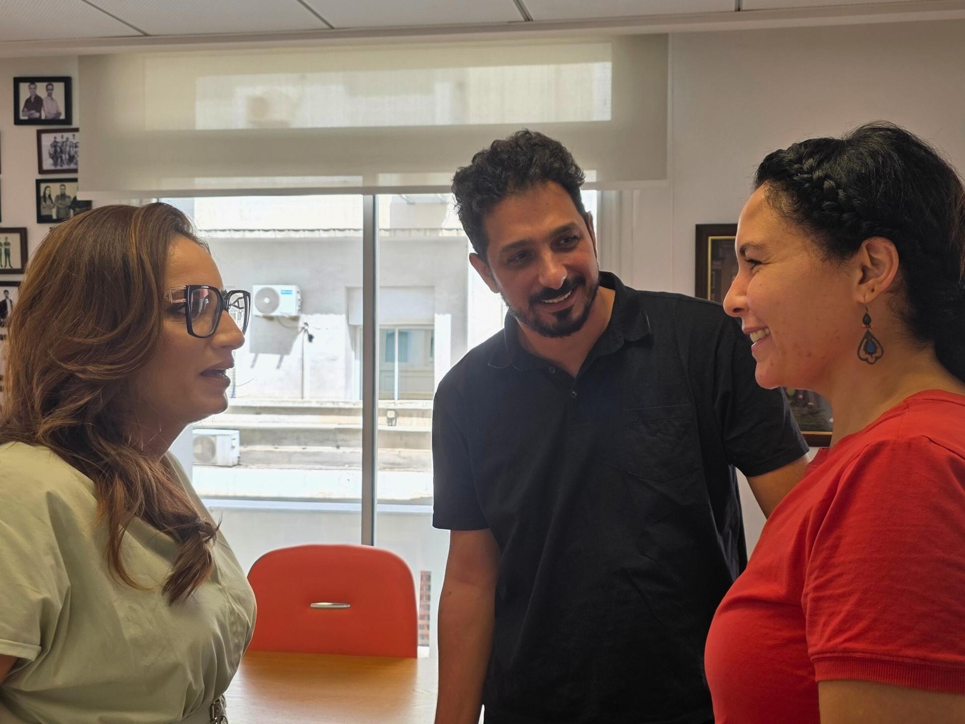 Three people talk in a bright room by a window; orange chairs nearby.