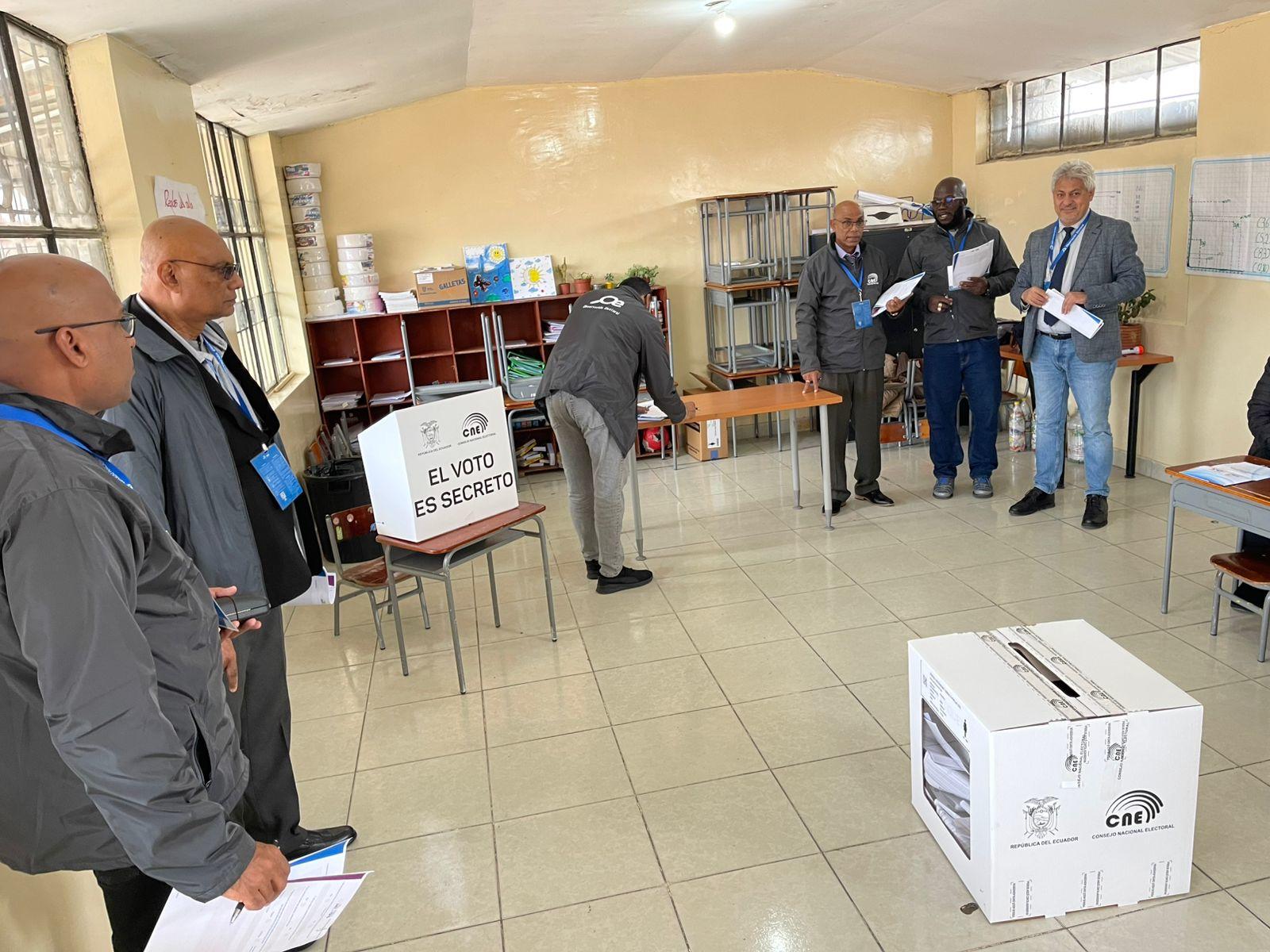 Photo: people at an indoor polling station around a ballot box.