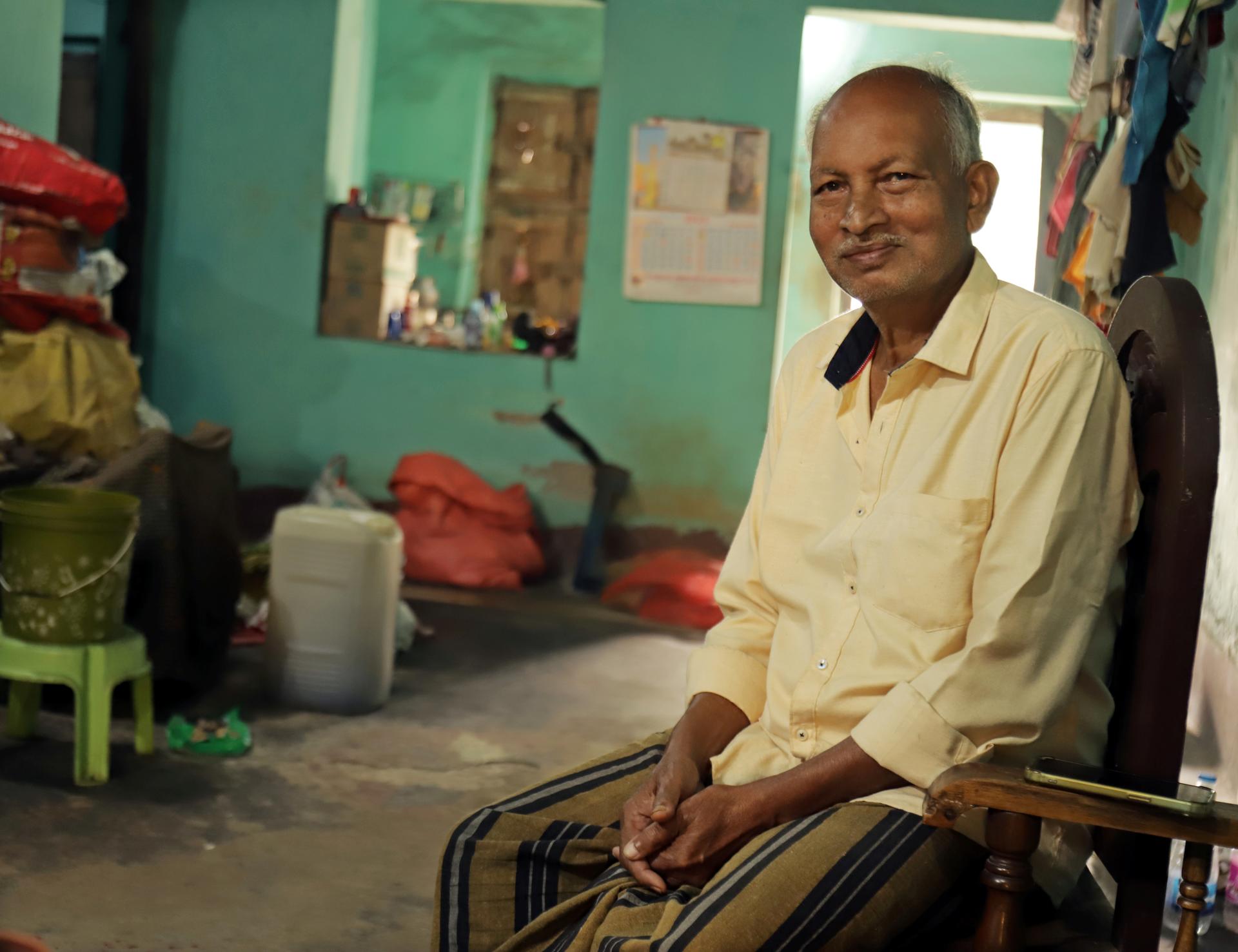 Man in pale yellow shirt sits on a wooden chair in a cluttered teal-walled room.