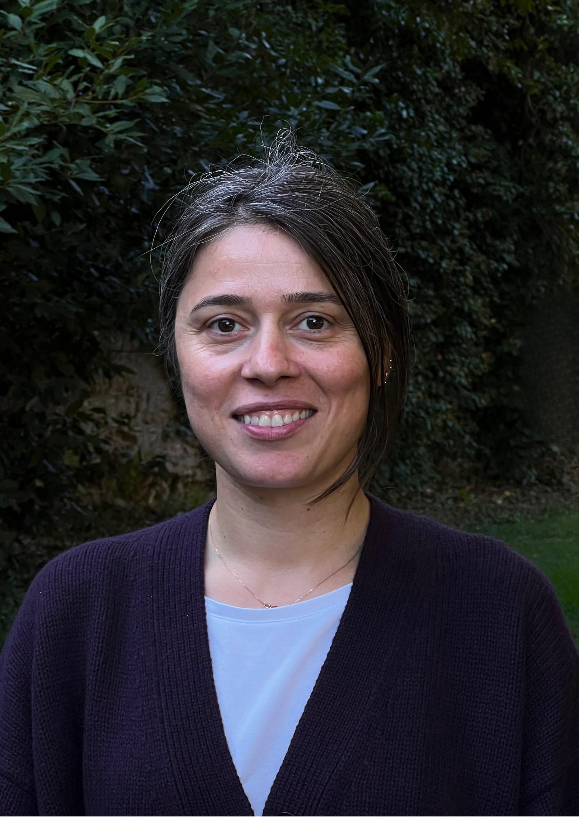 Person outdoors in front of green foliage, wearing a dark purple cardigan over a light blue top.