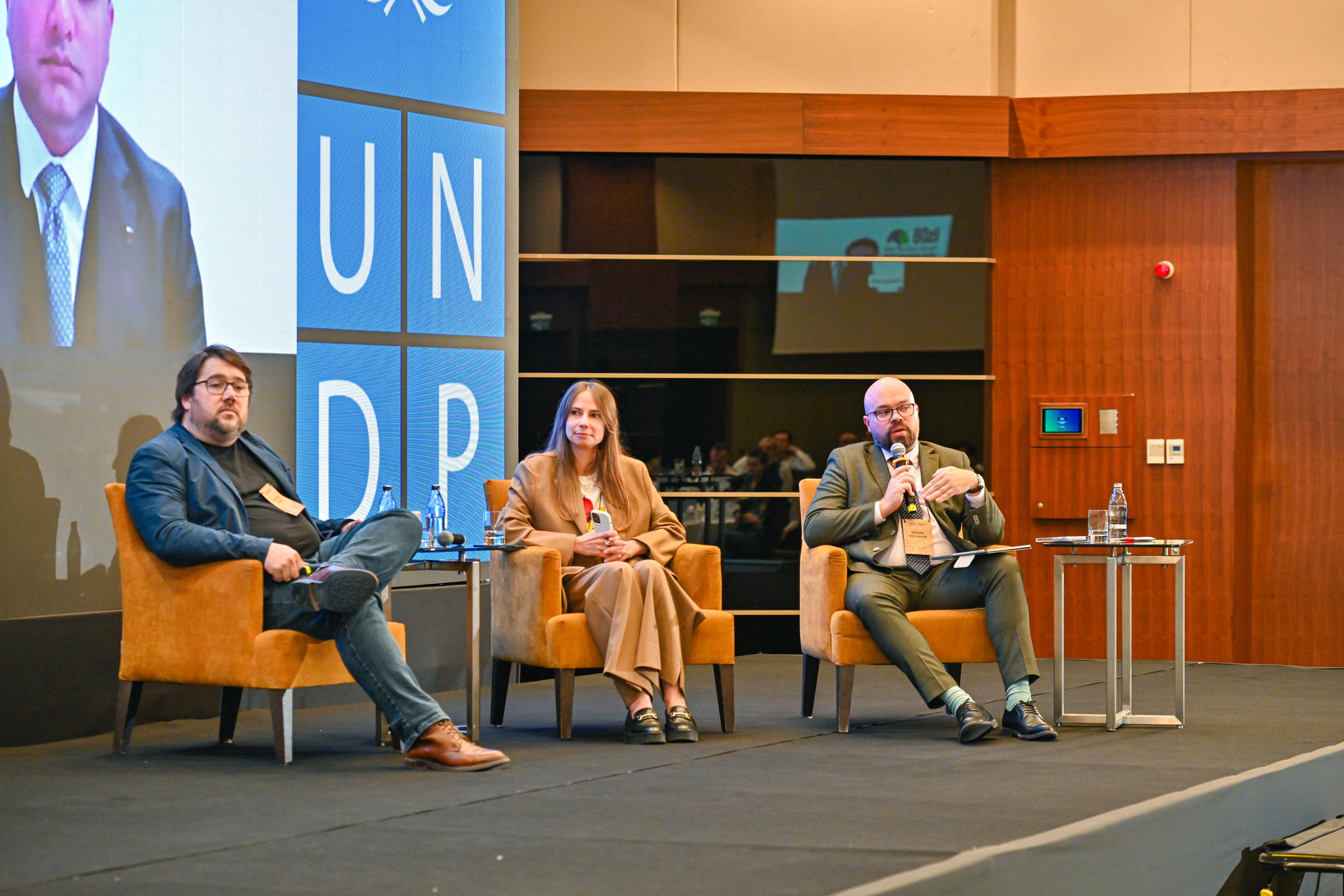 Photo: Four panelists in orange chairs on stage with UNDP branding; screen shows a speaker.