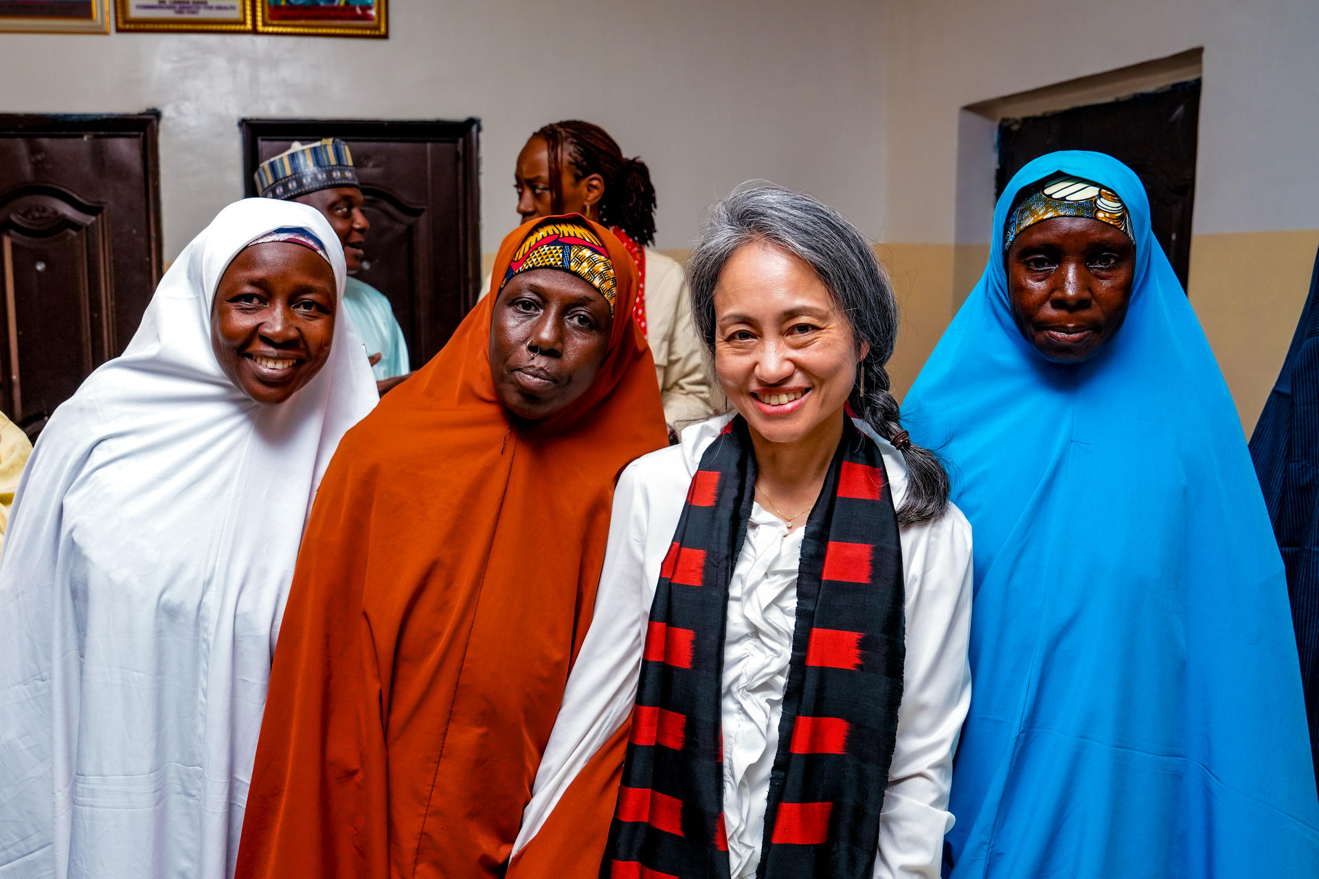 Group of five people in colorful robes standing indoors; front person wears a patterned scarf.