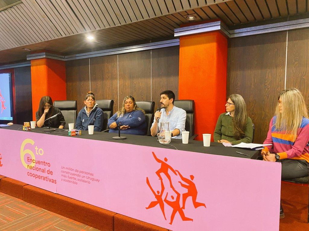 Panel of speakers seated behind a bright pink desk with a red running-figure logo banner.