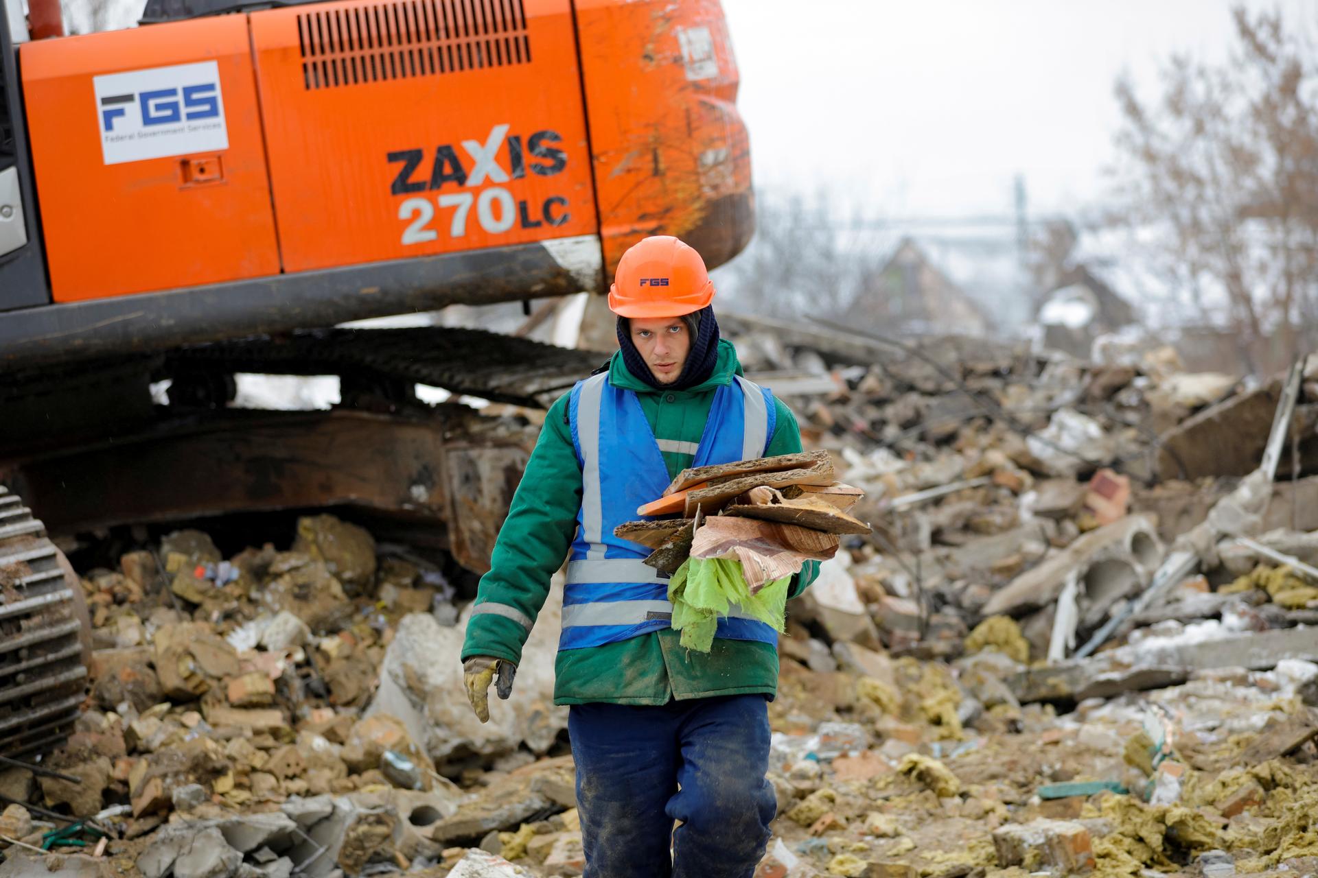 Worker in orange hard hat and teal jacket with clipboard amid rubble, orange excavator behind.