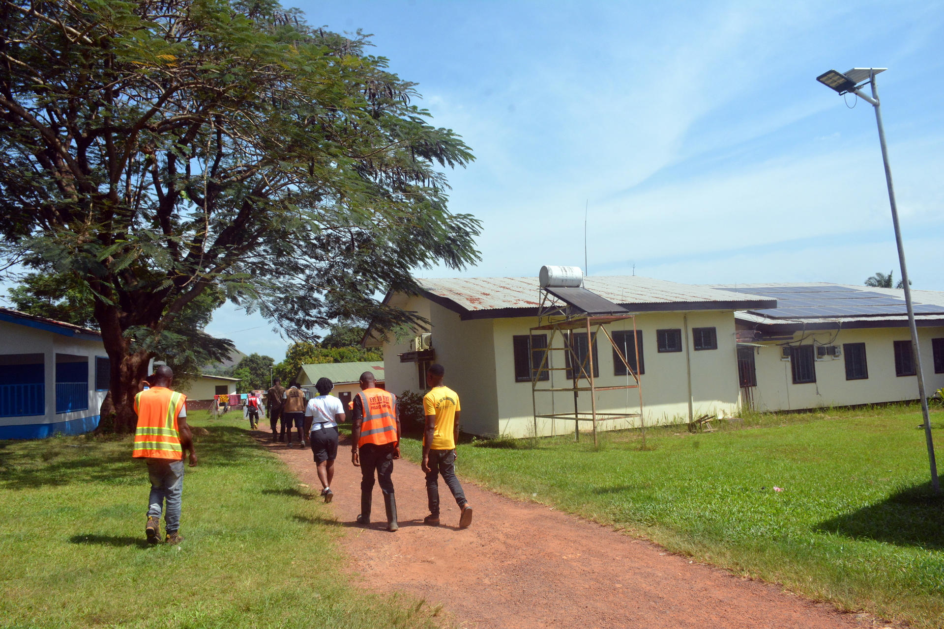 People in orange vests walk along a dirt path toward yellow buildings under a blue sky.