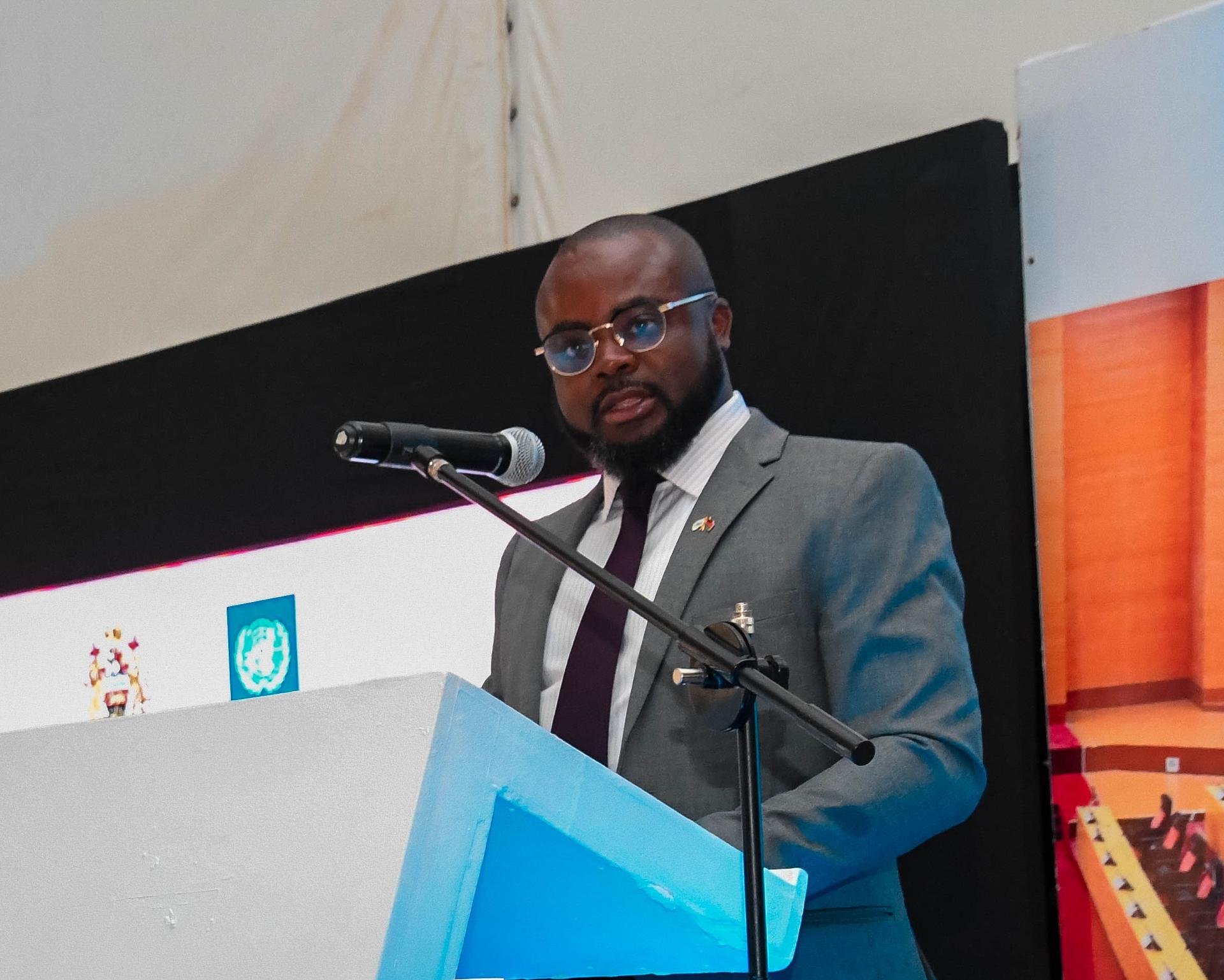 A man in a suit giving a speech at a blue podium with a microphone; laptop with logos nearby.