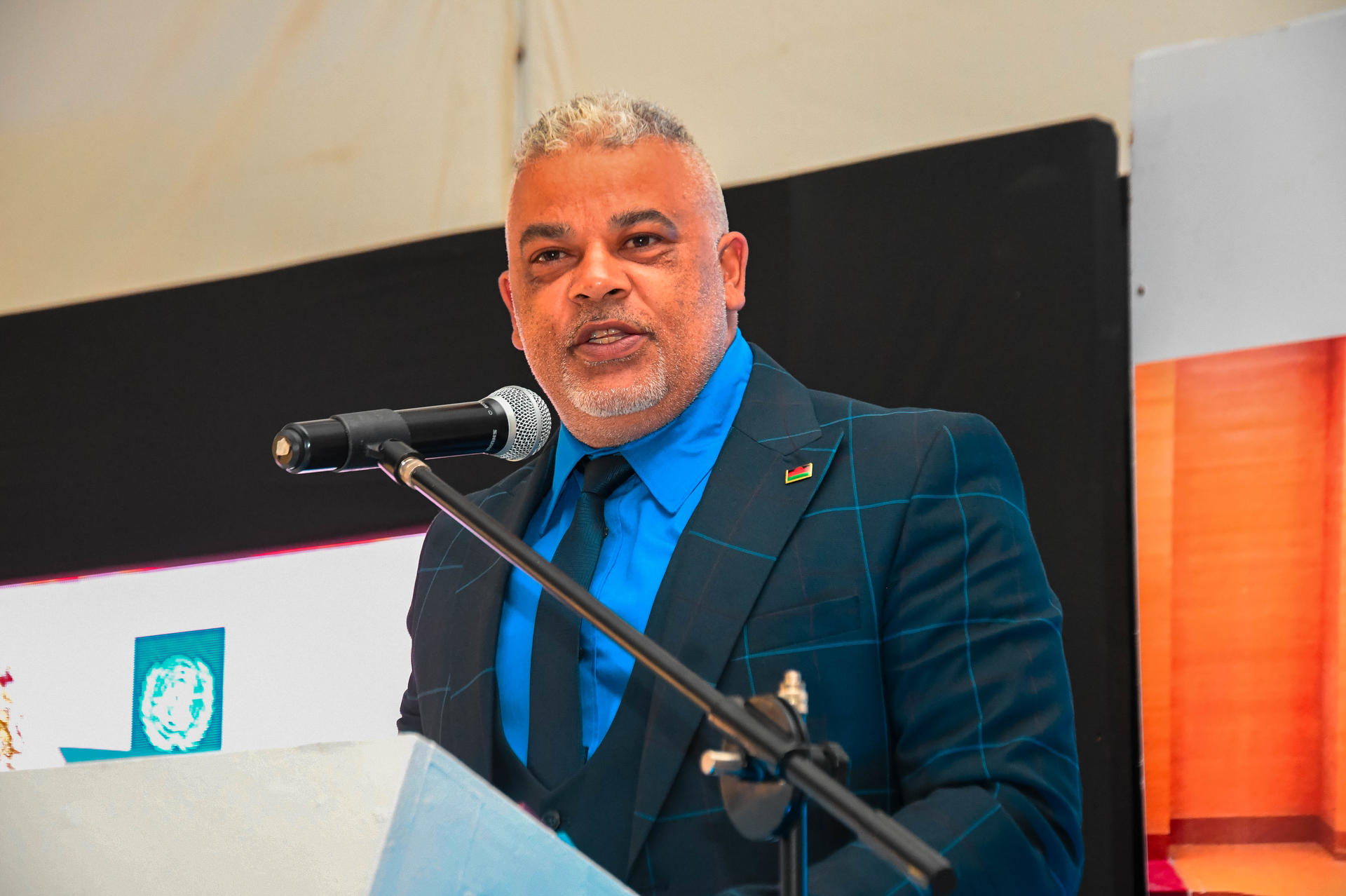Man in blue suit and light blue shirt speaks at a podium with microphone on stage.