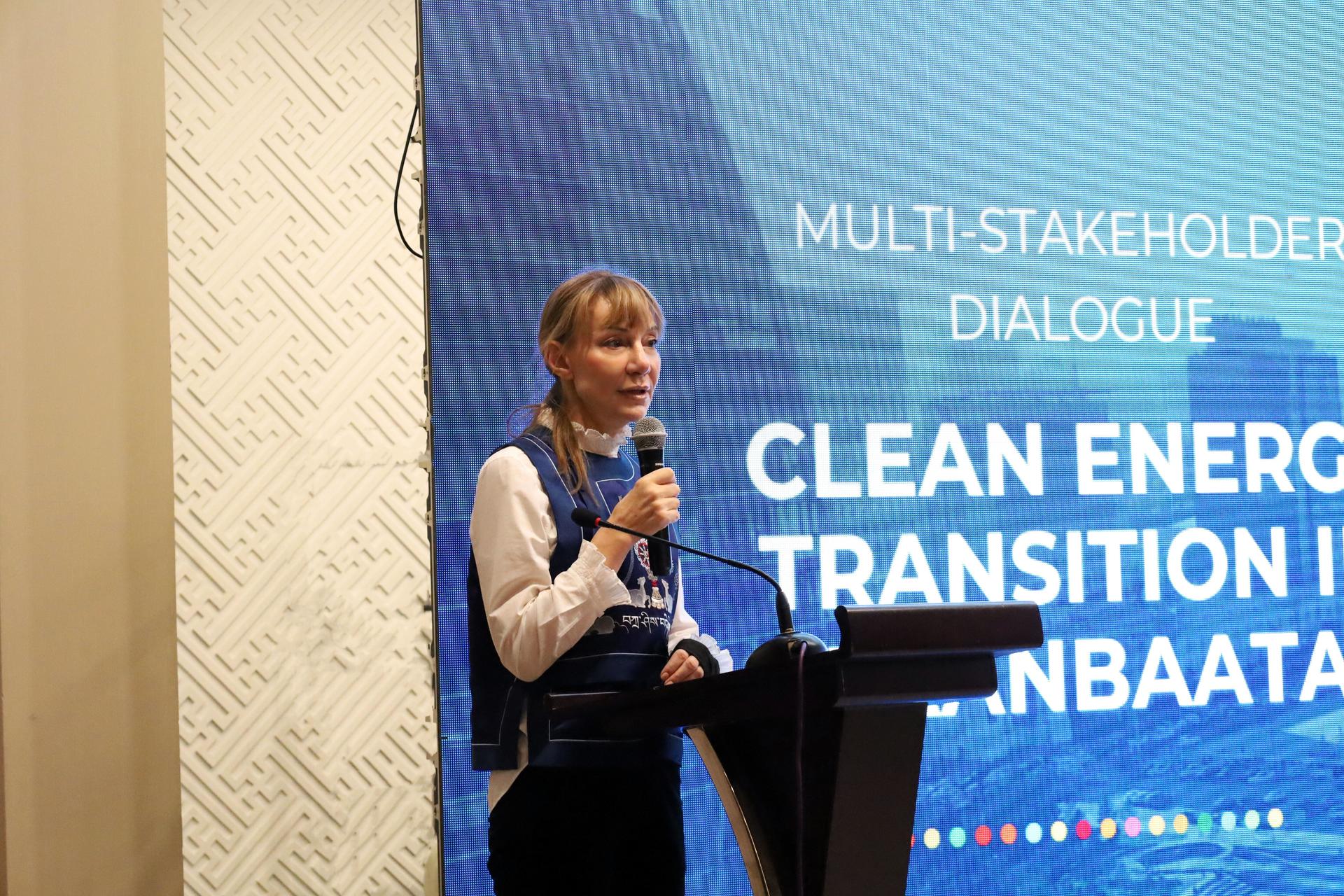 Female speaker at podium with a blue conference screen about clean energy transition.